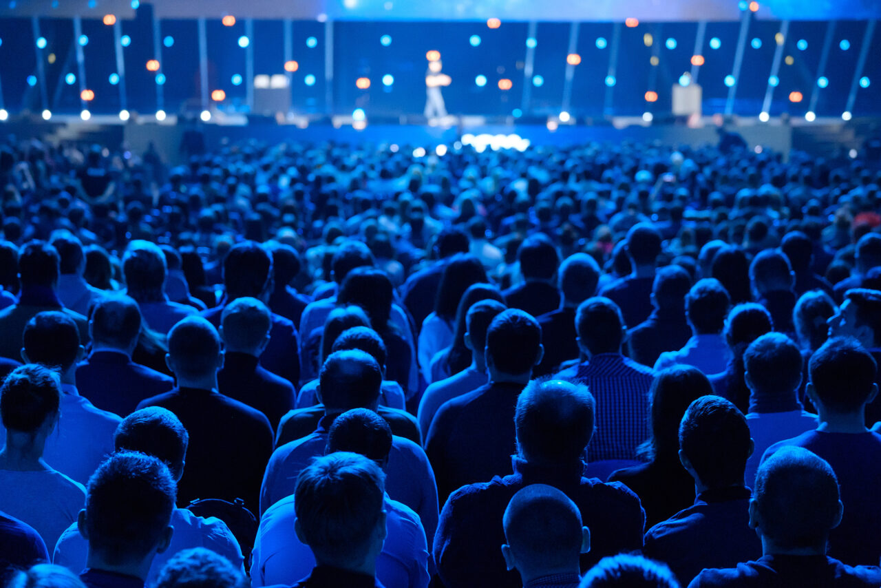 A large audience sits facing a stage at a conference, illuminated by blue lighting.