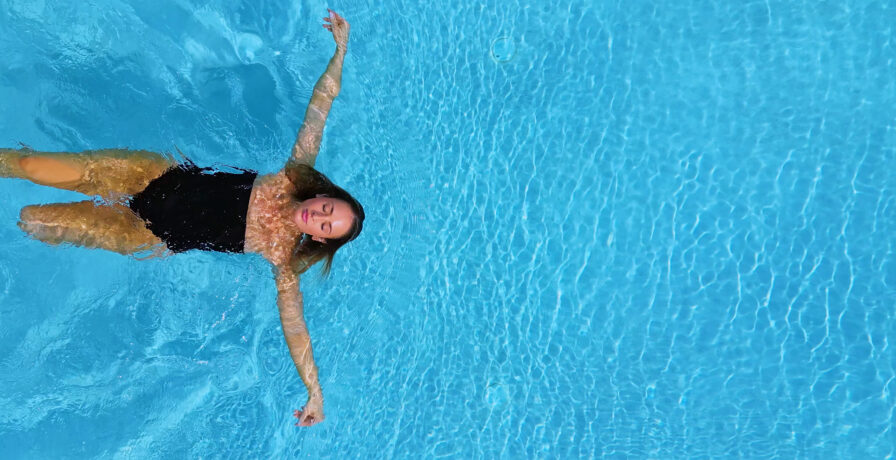 Person floating on their back in a blue swimming pool, wearing a black swimsuit.