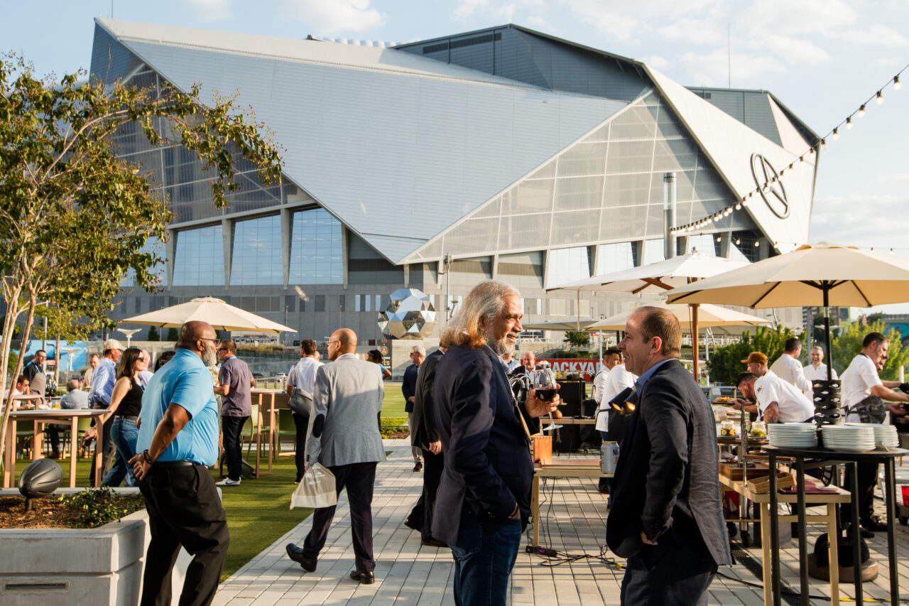 People socialize outdoors at an event with food stands and tables. A large, modern building serves as the backdrop. Sunny weather and string lights create a festive atmosphere.