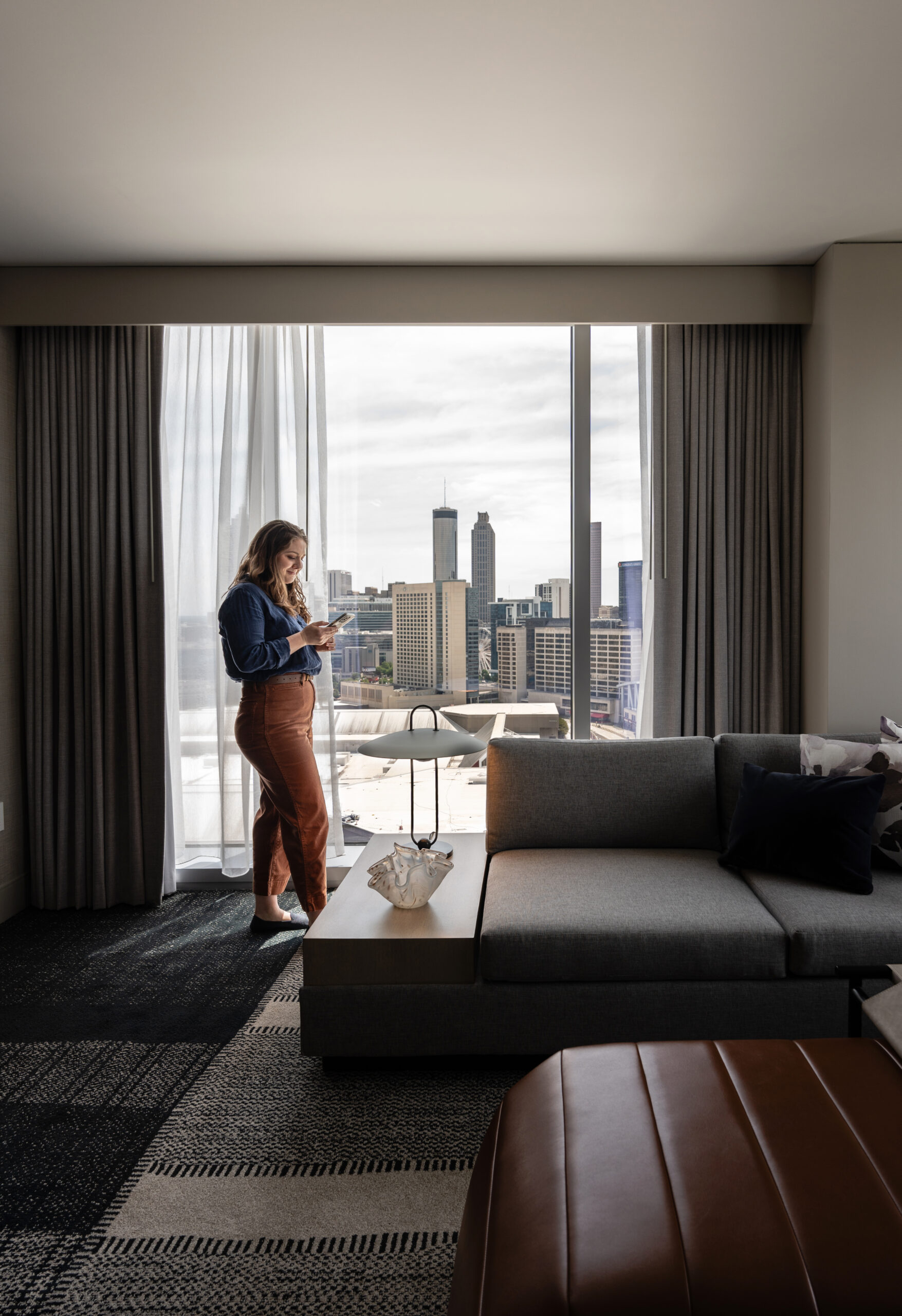 A person stands near a large window in the living room, absorbed in their smartphone. The window offers a stunning cityscape view with towering buildings. The cozy room features a couch, table, and elegant curtains, creating the perfect suite for comfort and relaxation.