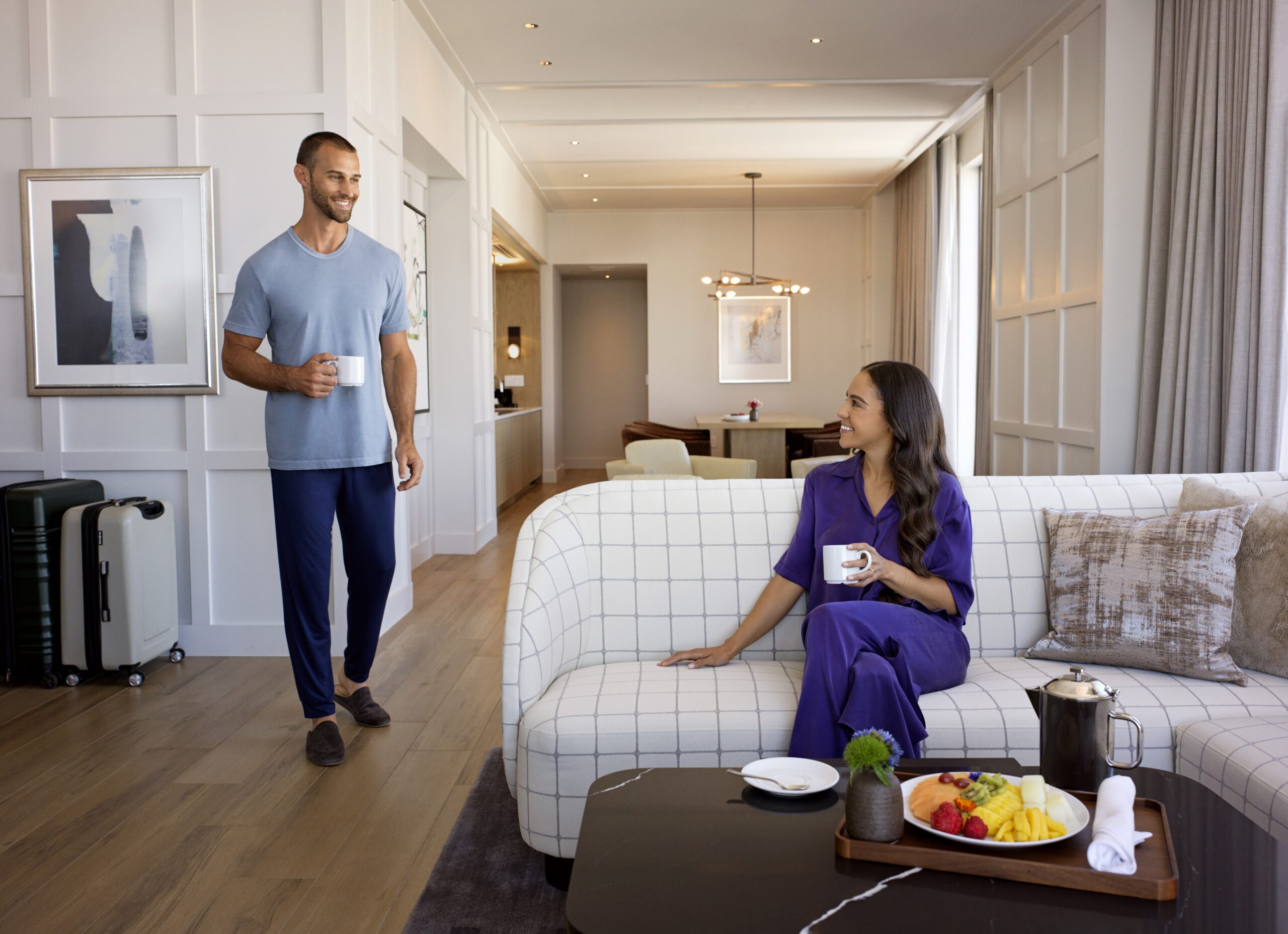 A man and woman in casual attire enjoy coffee in a modern, spacious suite with a tray of fruit on a table. The room features minimalistic decor and a large window with curtains, offering an inviting atmosphere.