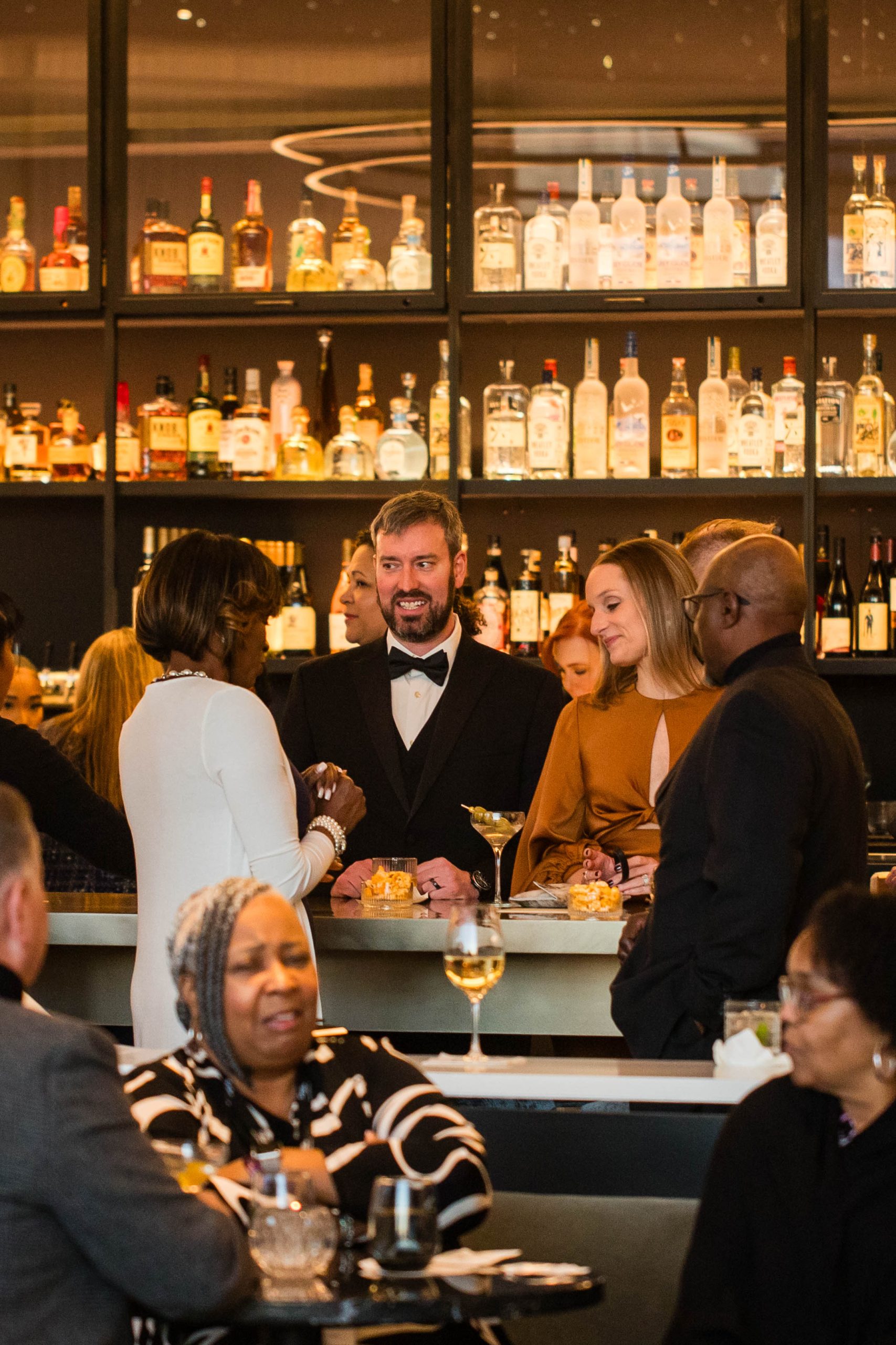 People dressed in formal attire socialize at a bar with a variety of liquor bottles displayed on shelves.