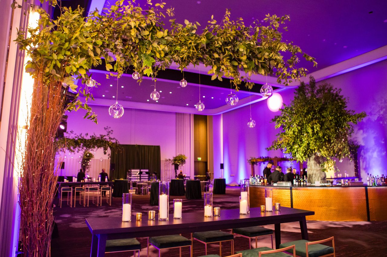 Decorated event space with a long table topped with candles, surrounded by chairs. Overhead greenery with hanging glass orbs. Illuminated in purple lighting, with a bar and additional seating.
