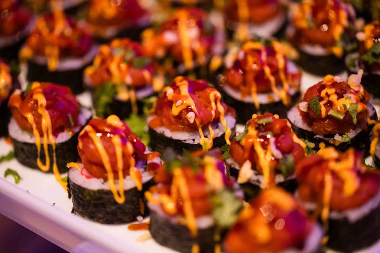 A platter of sushi rolls topped with raw fish, bright orange sauce, and garnished with herbs sits gracefully on a white tray, perfect for an intimate private dining experience.