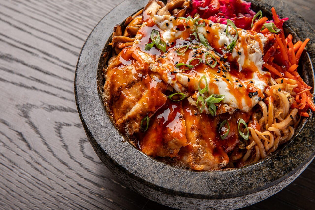 A stone bowl filled with noodles, grilled chicken, fried egg, shredded vegetables, and sauce, garnished with green onions and sesame seeds on a wooden table.
