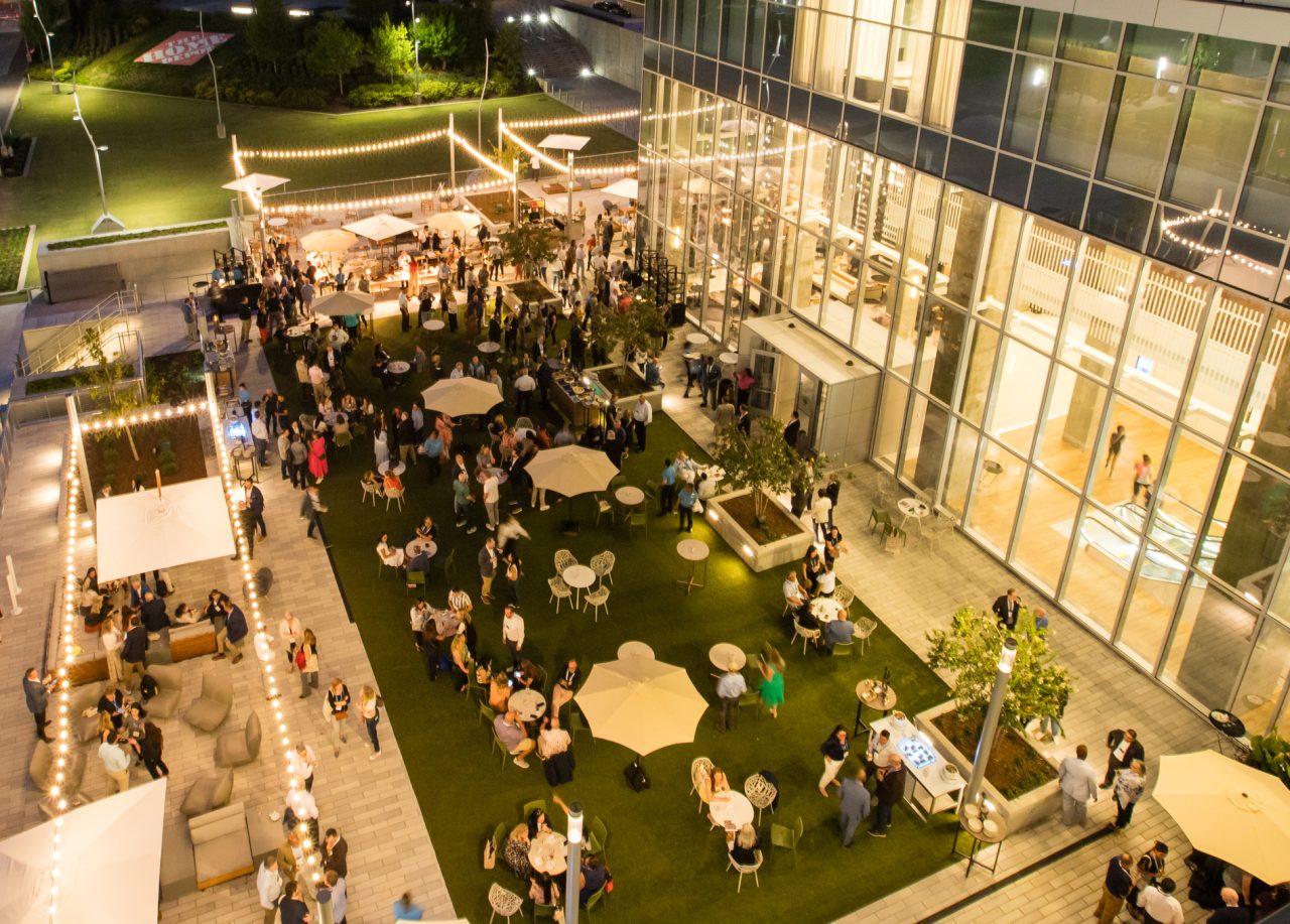Aerial view of an outdoor evening event with people gathered around tables on a green lawn, surrounded by string lights and adjacent to a modern glass building.