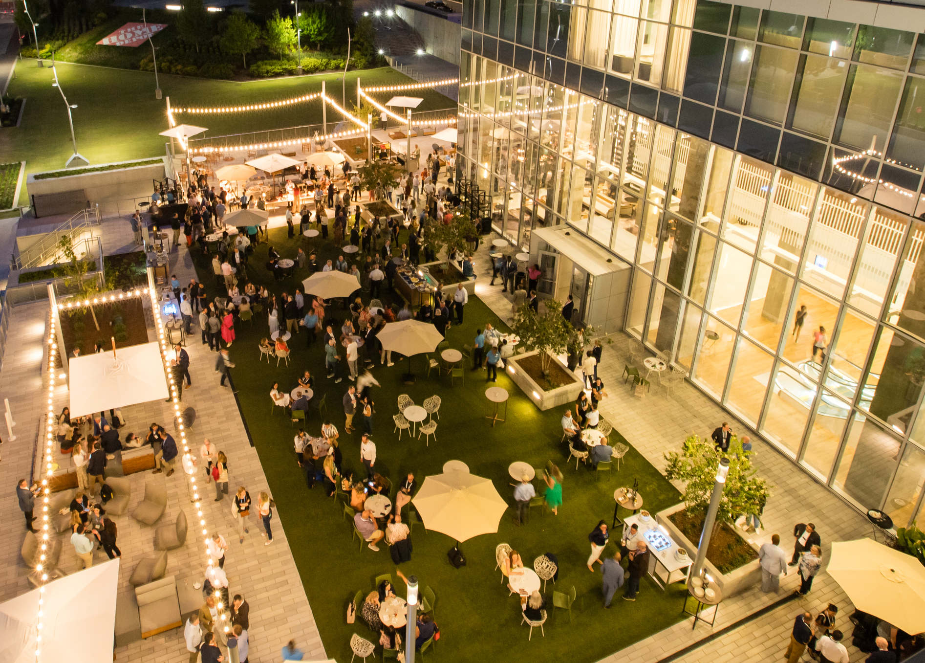Aerial view of an outdoor evening event with people gathered around tables on a green lawn, surrounded by string lights and adjacent to a modern glass building.