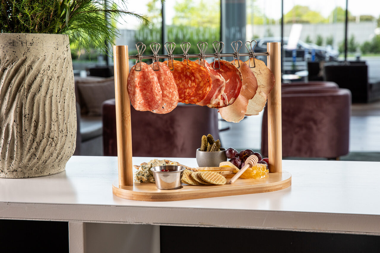 A charcuterie board featuring assorted sliced meats hanging on hooks, with crackers, cheese, grapes, pickles, and dipping sauce arranged on a wooden tray.