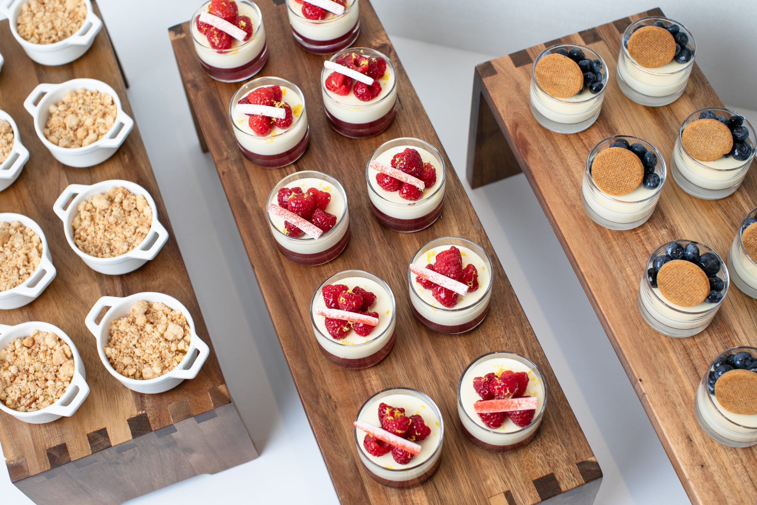 An assortment of small desserts is artistically arranged on wooden stands for an intimate private dining experience, featuring berry-topped parfaits, crumbles in white ramekins, and delights garnished with cinnamon cookies and blueberries.