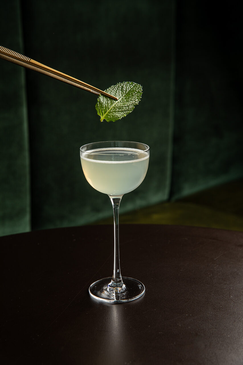 A hand holding tongs places a mint leaf garnish on a pale yellow cocktail in a coupe glass, set on a dark table with a green background.