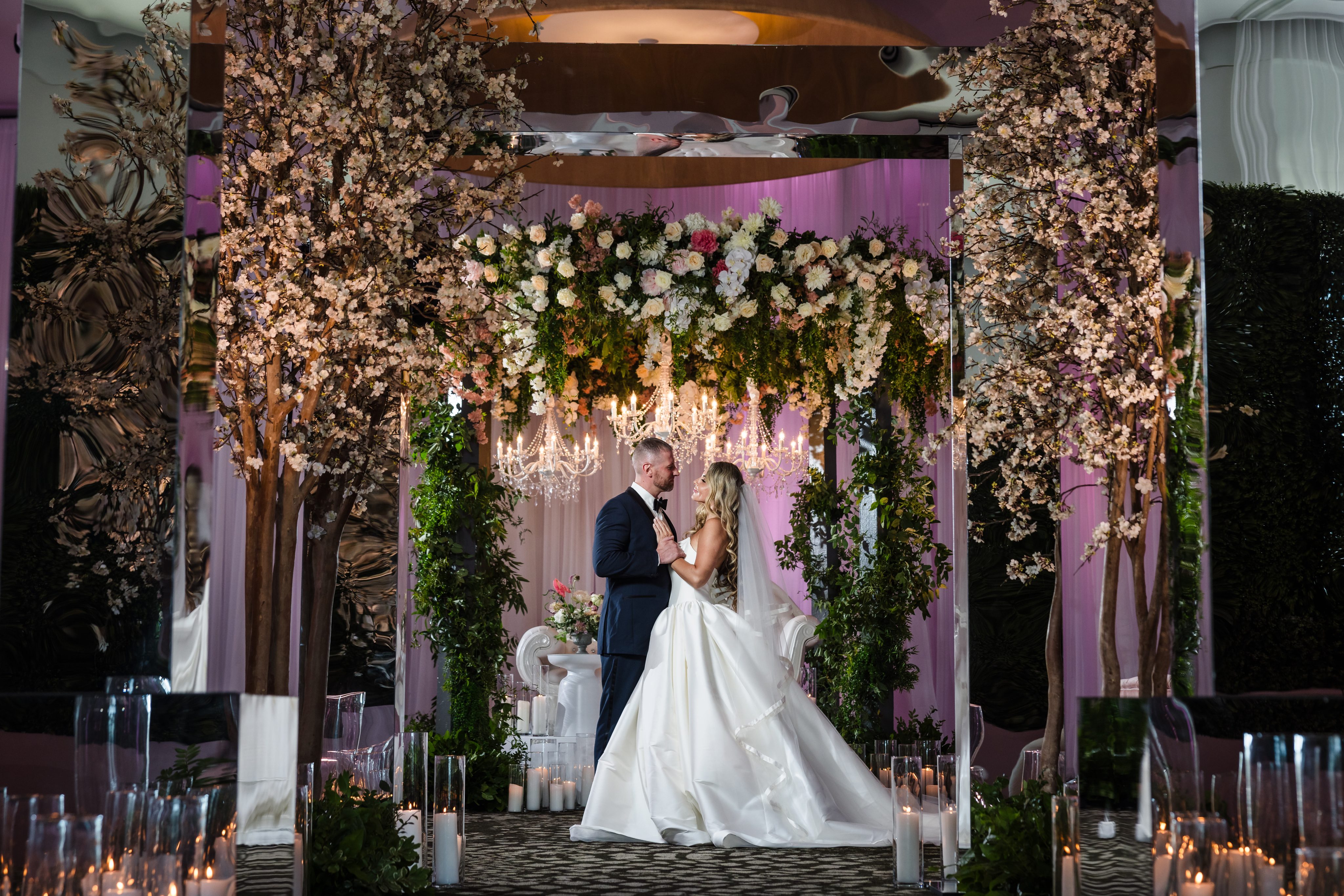 Bride and groom stand under a floral arch surrounded by candles and greenery. A chandelier hangs above them, providing illumination.