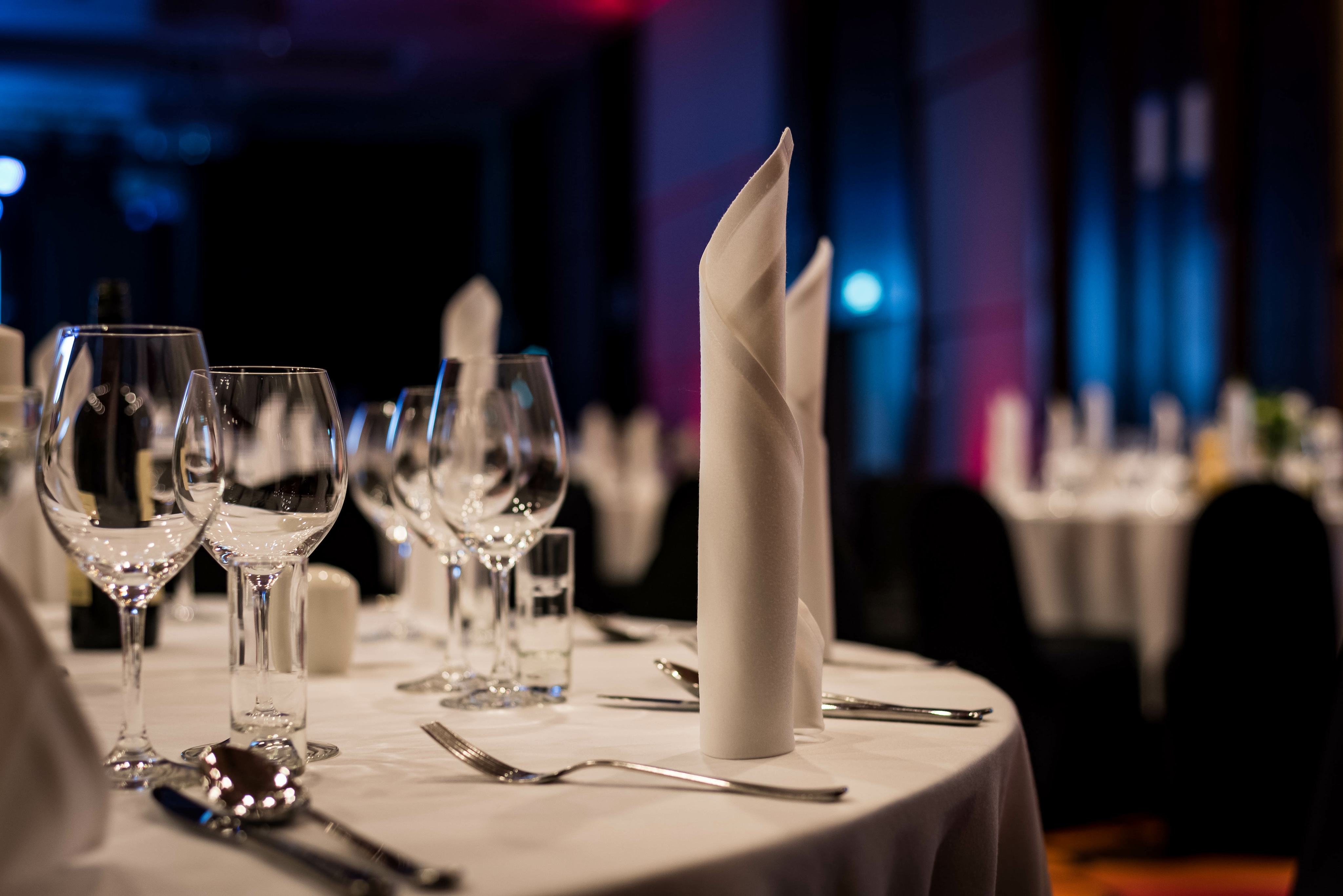 A formally set dining table with folded napkins, wine glasses, water glasses, and silverware. Dimly lit room with other tables visible in the background.