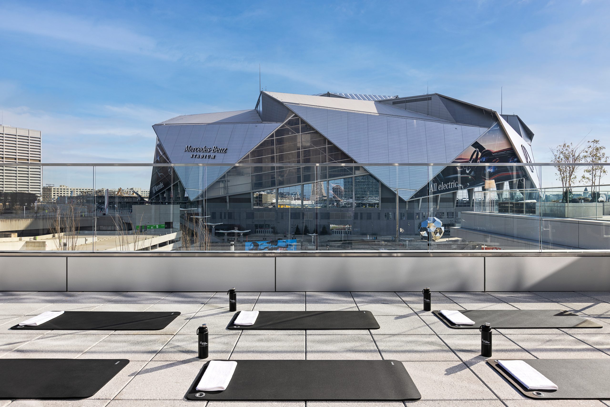 Outdoor rooftop yoga mats and water bottles are set up in front of a large, modern stadium building under a clear blue sky.