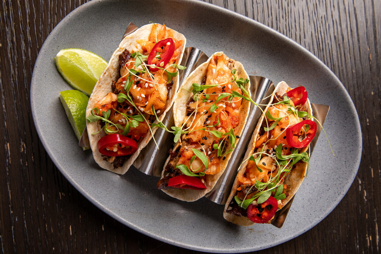 Three tacos filled with vegetables, microgreens, and sliced red chili peppers are arranged on a gray oval plate with lime wedges on the side.