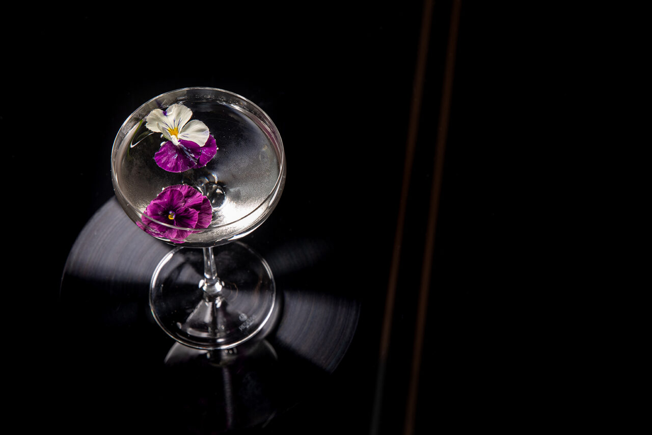 A clear cocktail in a coupe glass garnished with two purple and one white edible flowers, set on a glossy black surface.