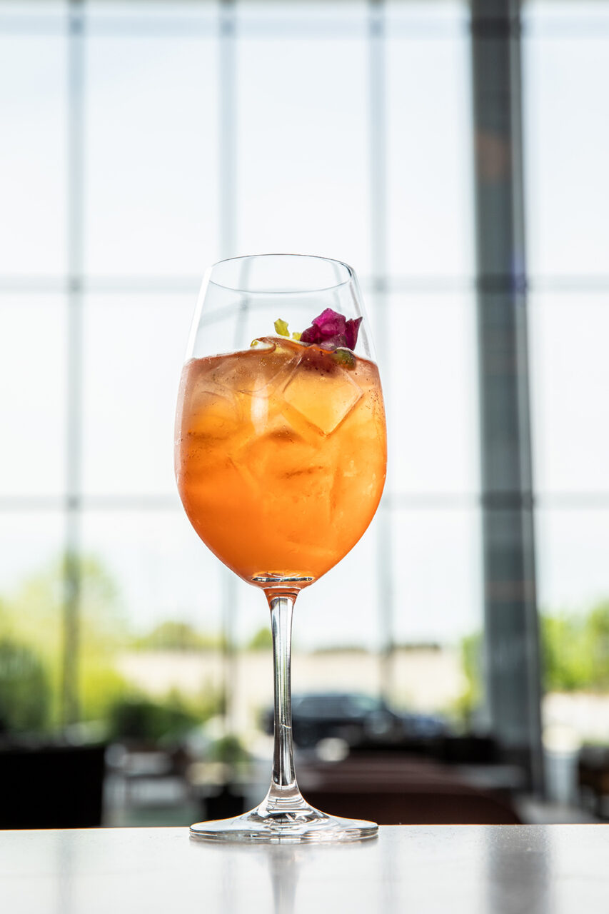 A stemmed glass filled with an orange-colored iced drink, garnished with a flower, is placed on a table in front of large windows.