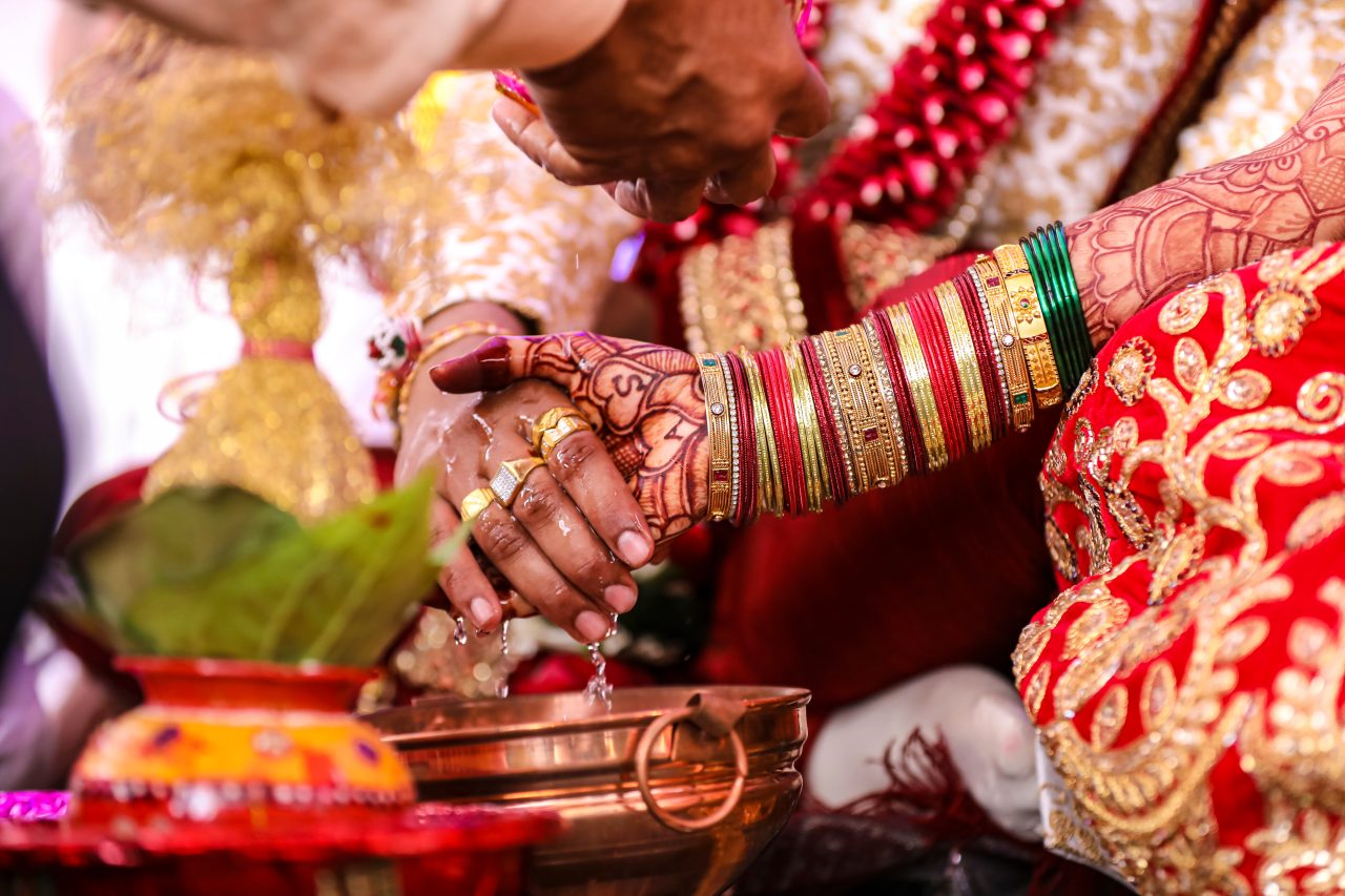 Close-up of a traditional wedding ritual with decorated hands holding each other over a copper vessel. Hands adorned with colorful bangles and intricate henna designs.