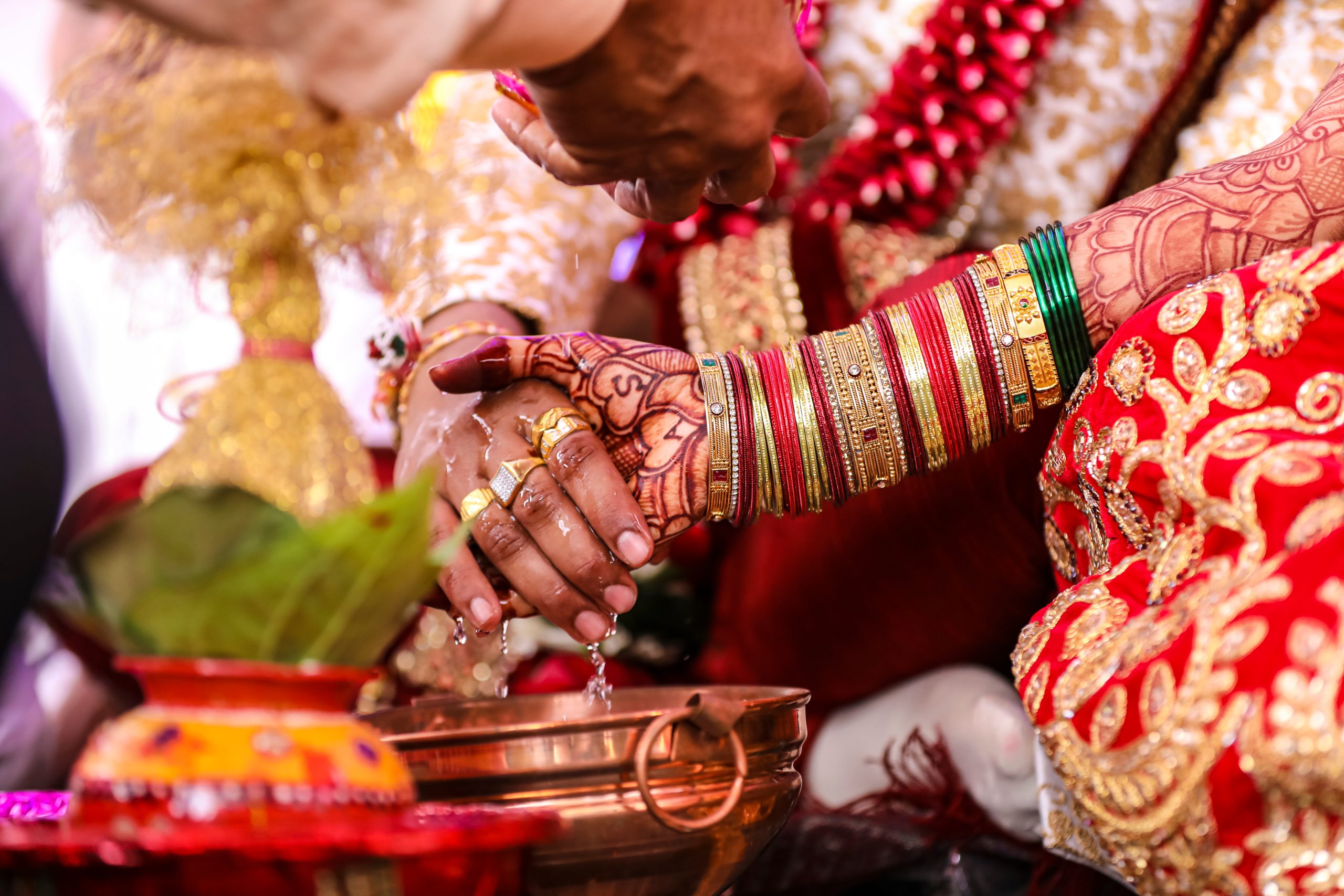 Close-up of a traditional wedding ritual with decorated hands holding each other over a copper vessel. Hands adorned with colorful bangles and intricate henna designs.