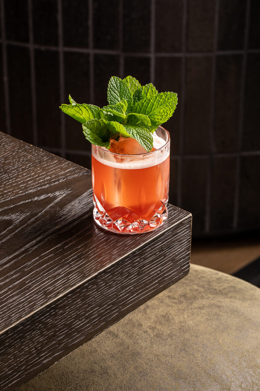 A glass of reddish cocktail garnished with fresh mint leaves and an orange peel on a wooden table.