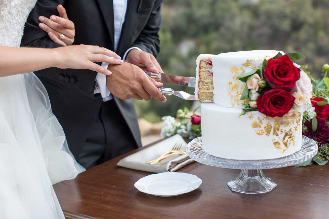 A couple cuts a two-tiered white cake with gold accents and red roses on a table. The cake slice reveals layered filling.