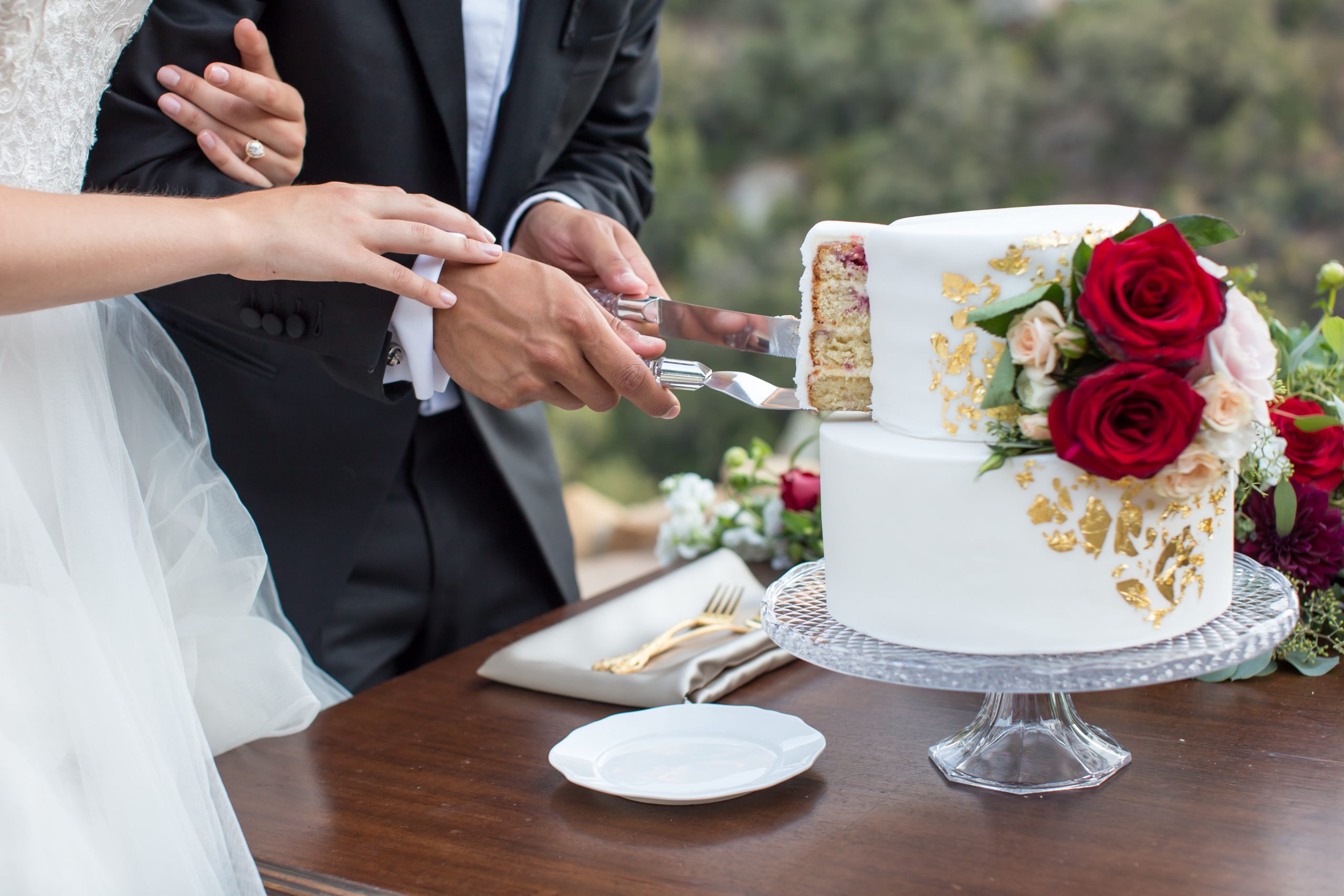 A couple cuts a two-tiered white cake with gold accents and red roses on a table. The cake slice reveals layered filling.