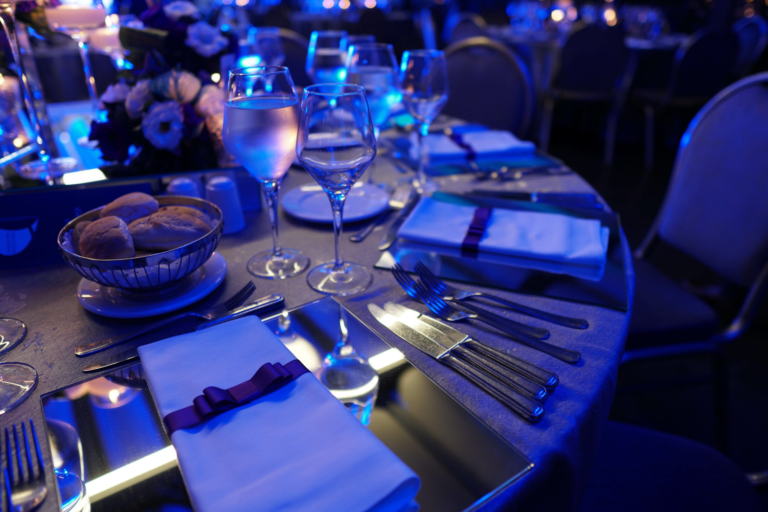 Elegant table setting with mirrored surfaces, wine glasses, cutlery, and napkins tied with purple ribbons under blue lighting; bread basket and floral centerpiece in the background.