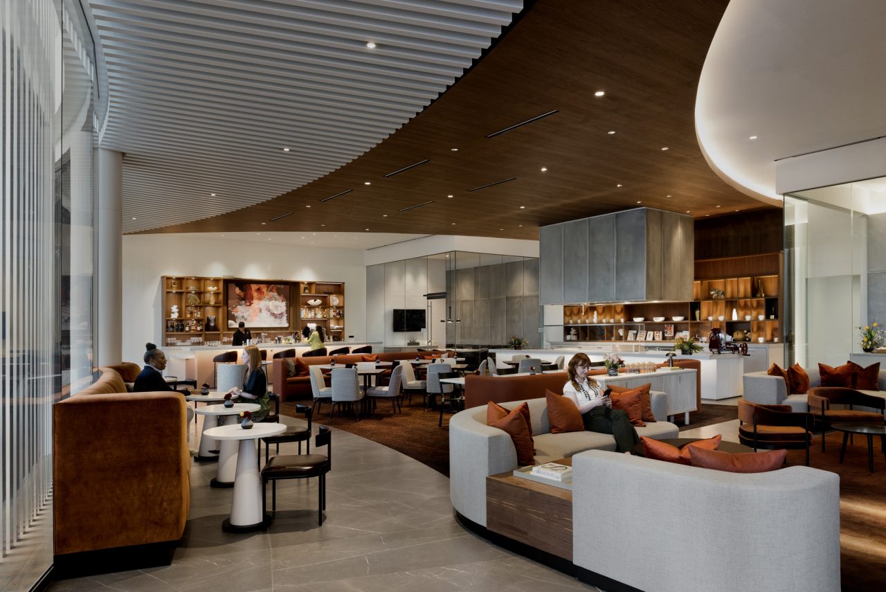 Spacious modern lounge with curved wooden ceiling, brown and beige furniture, and people sitting at tables. Large windows on the left allow natural light to enter.