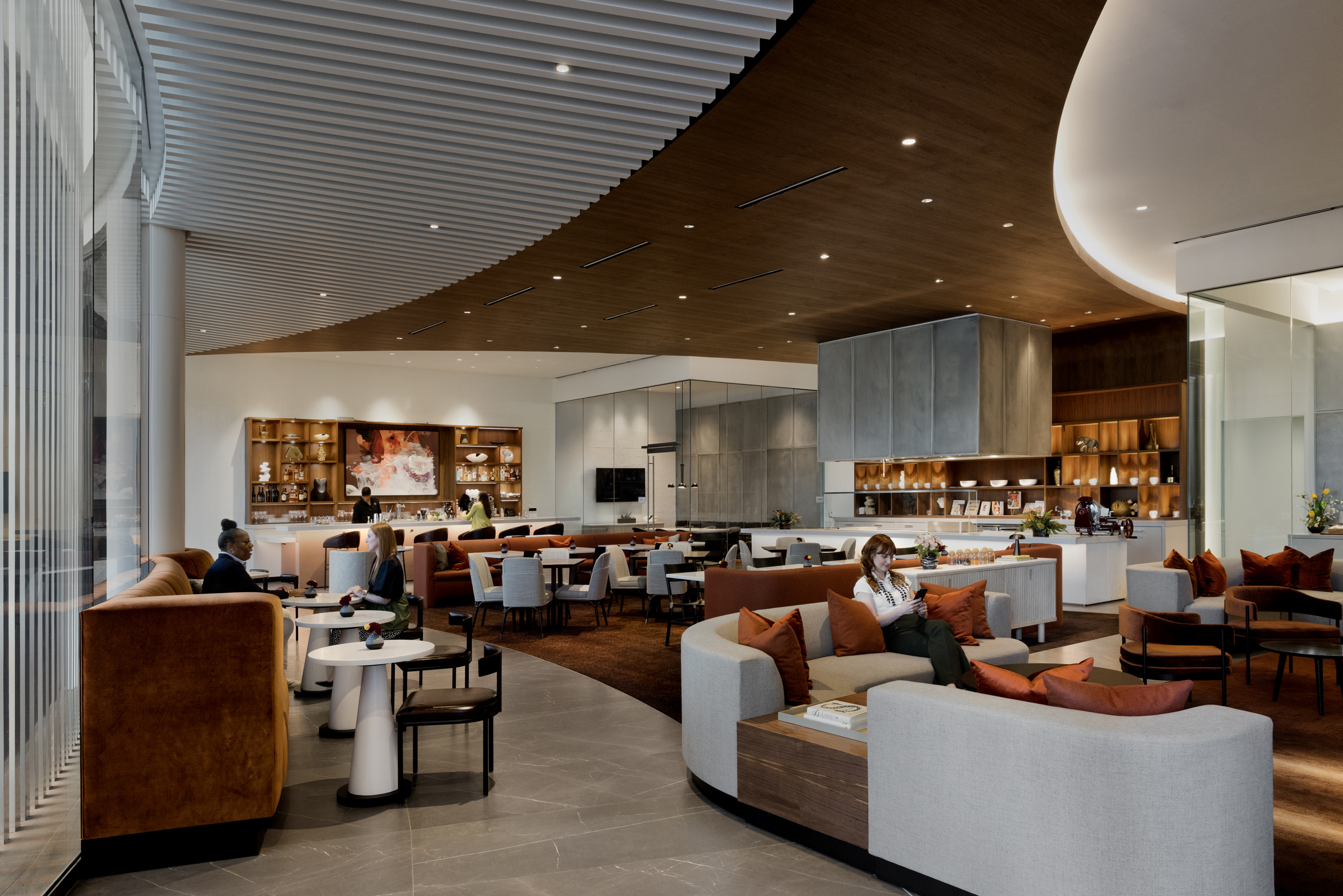 Spacious modern lounge with curved wooden ceiling, brown and beige furniture, and people sitting at tables. Large windows on the left allow natural light to enter.