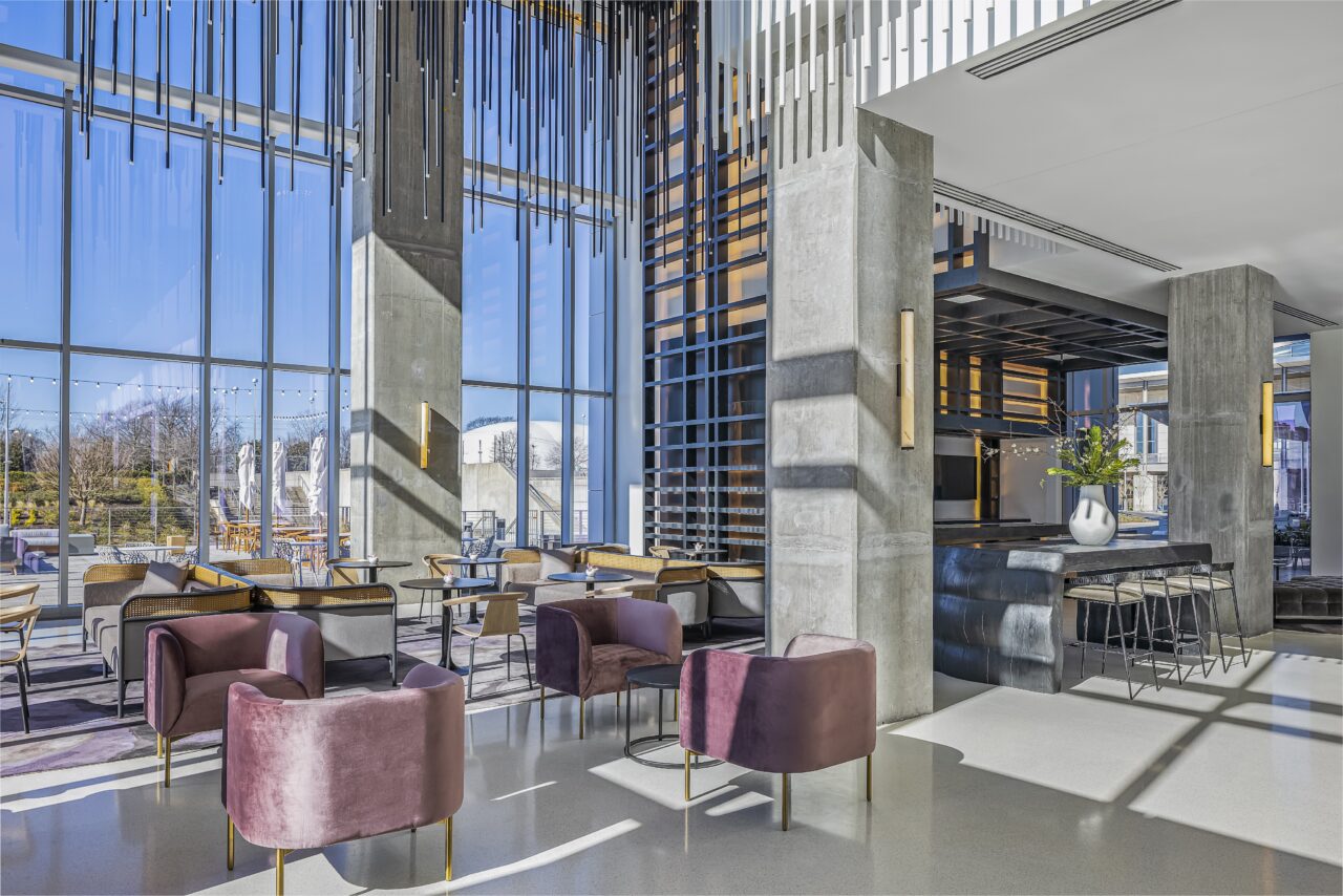 Modern hotel lobby with large windows, purple and gray chairs, a sleek reception desk, and hanging decorative elements. Sunlight streams in, illuminating the space.