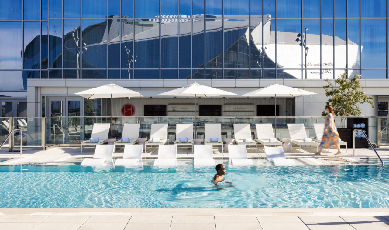 A person swims in a clear outdoor pool with white lounge chairs and umbrellas; a woman in a dress walks nearby. A glass building reflects a stadium across the street—ask about available AAA Discount for your stay.
