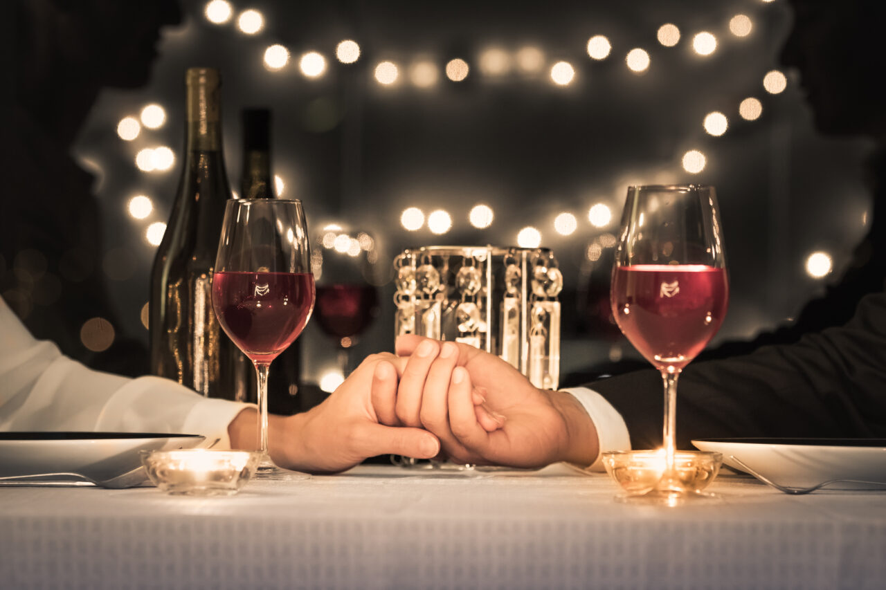Two people sit across from each other at a table, holding hands. Two glasses of red wine, bottles, and candles are on the table with string lights in the background—perfect for a cozy date night or romantic evening.