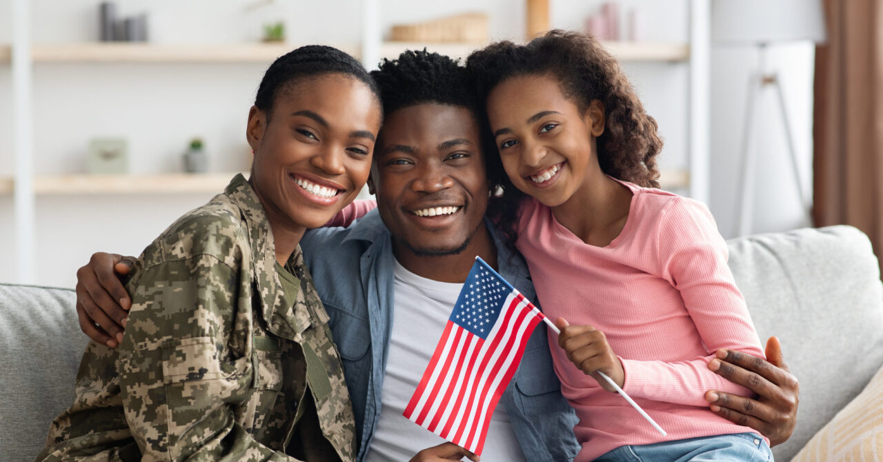 Three people sit on a couch smiling; the woman wears a military uniform, while the man and girl hold small American flags, proudly representing their bond and eligibility for the Military Family Rate.