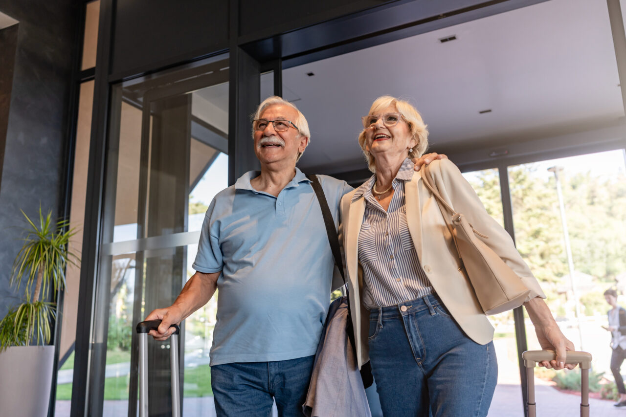An older man and woman with suitcases enter a building together, smiling and walking side by side near large glass doors.