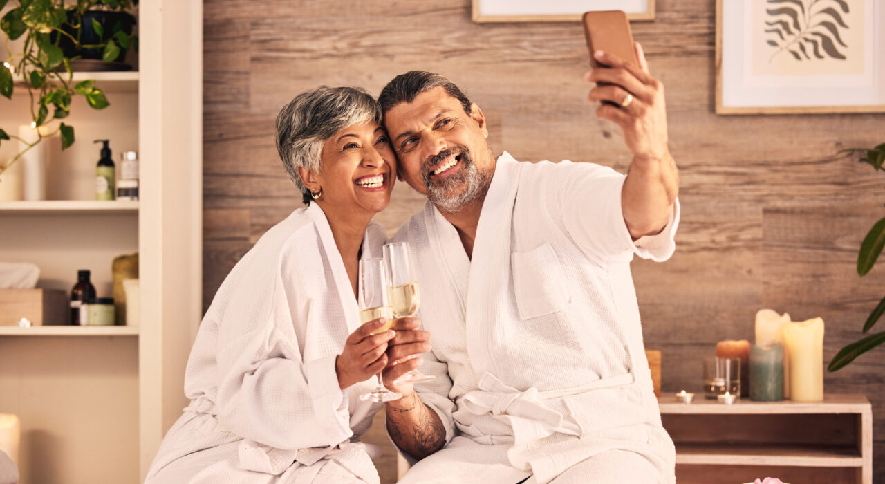 An older couple in white bathrobes sits together, smiling and taking a selfie with champagne glasses—enjoying a cozy, spa-like staycation that lets them escape the ordinary.