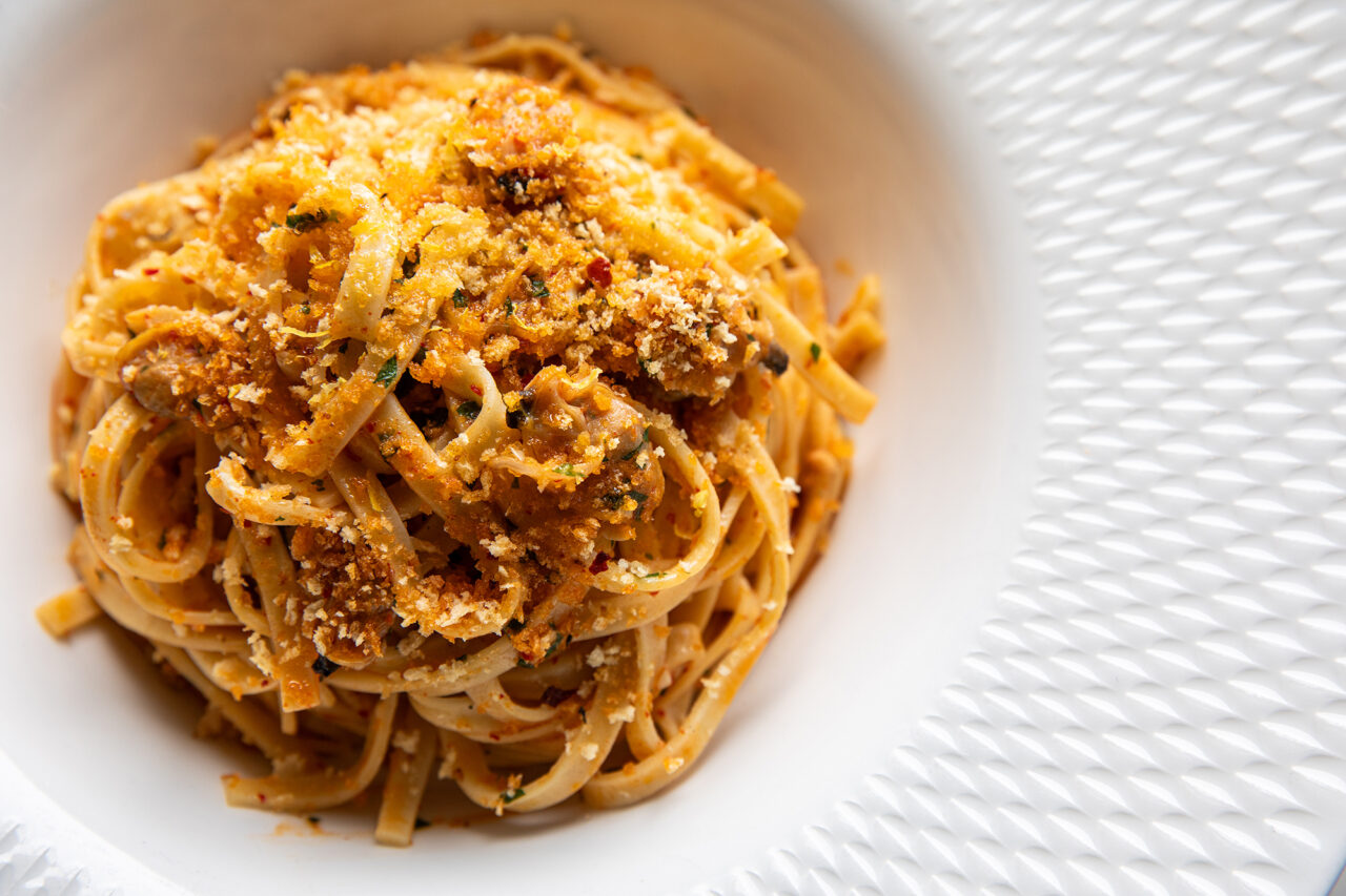 A close-up of a bowl of linguine pasta topped with a tomato-based meat sauce and grated cheese, served on a textured white plate.
