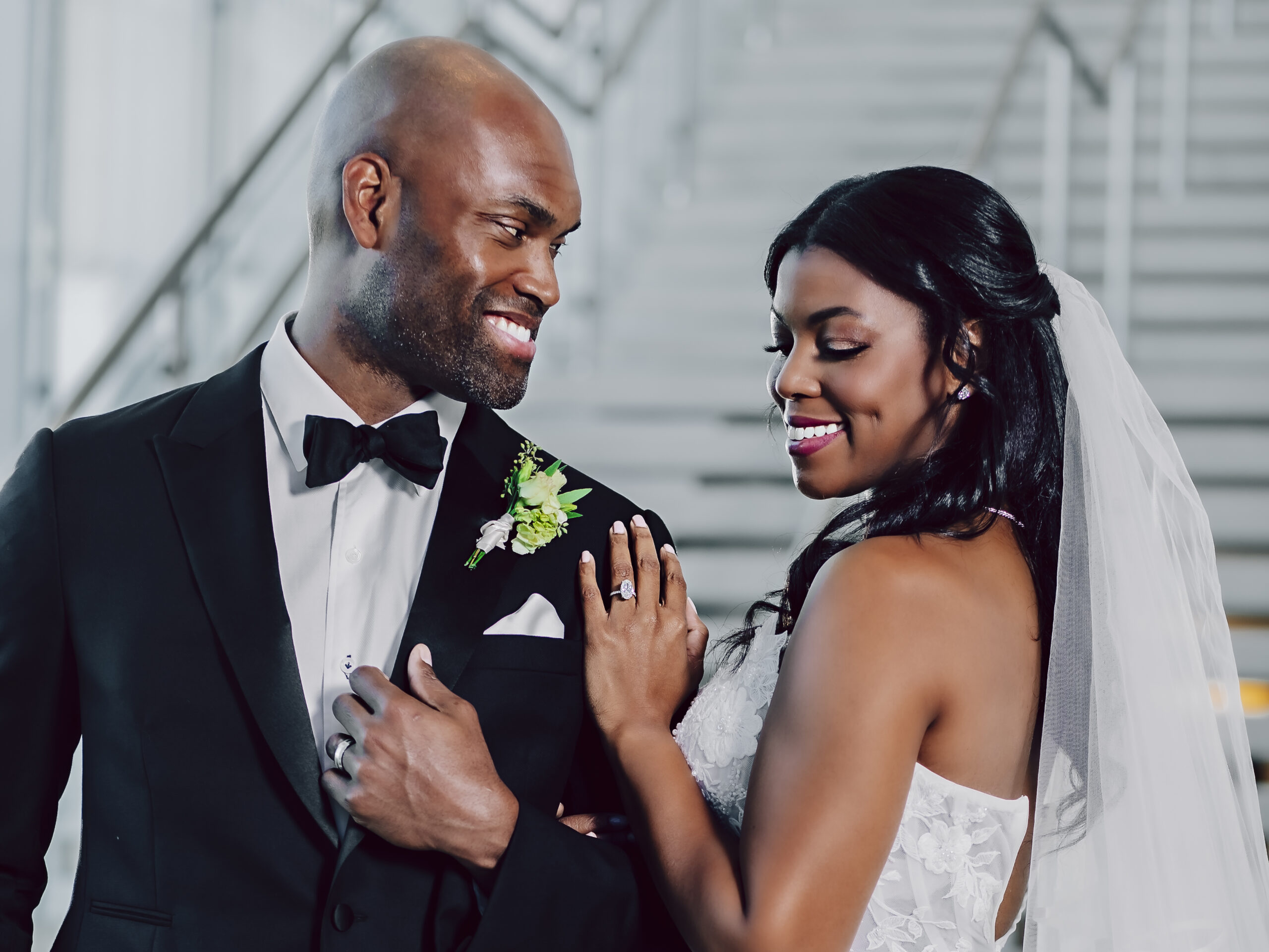 A bride in a white dress and veil stands beside a groom in a black tuxedo, both smiling and holding each other's arms in front of a staircase.