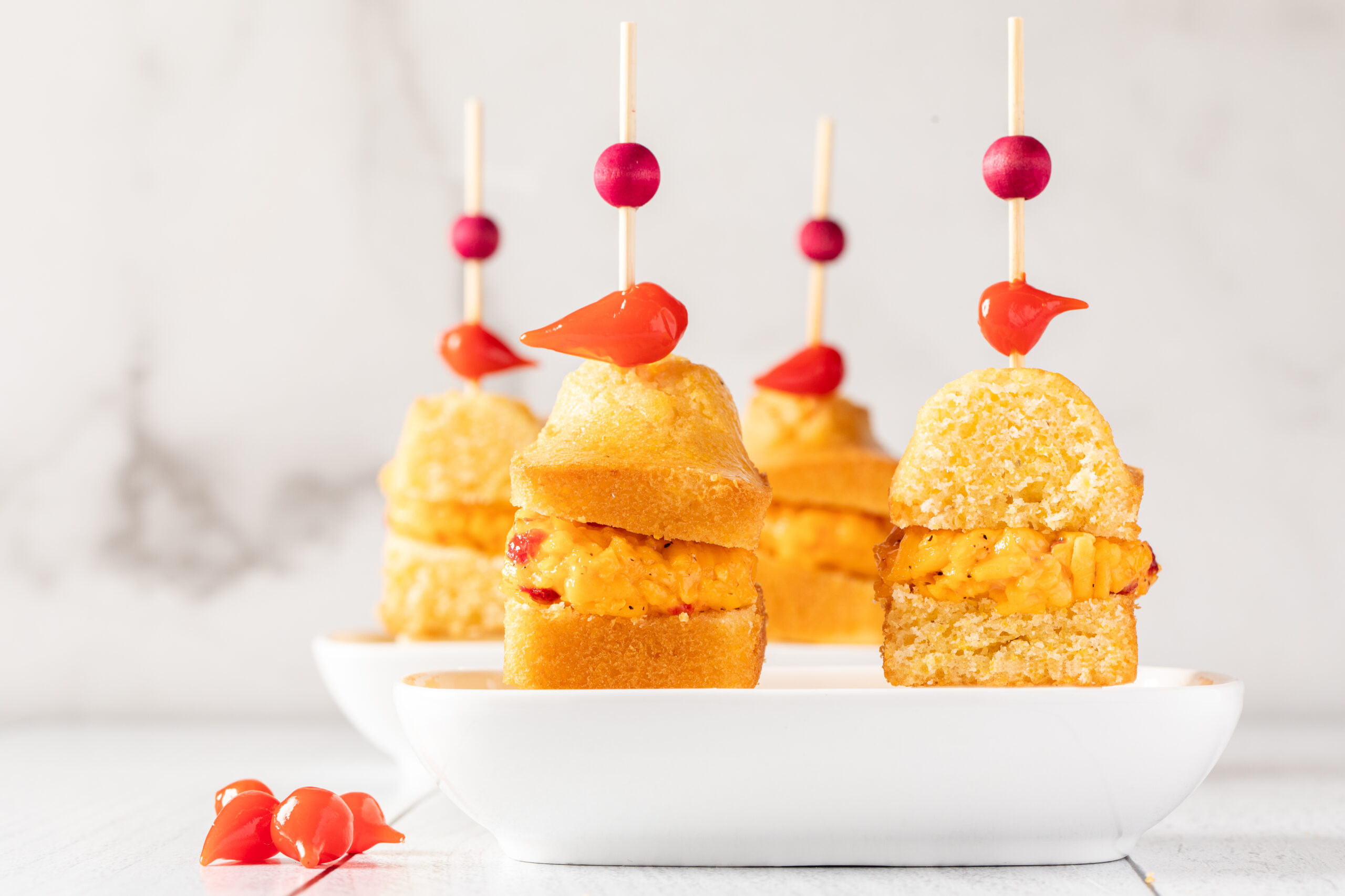 Three mini cornbread sandwiches filled with a yellow spread are secured with skewers, each topped with a red pepper and bead, presented on a white dish.