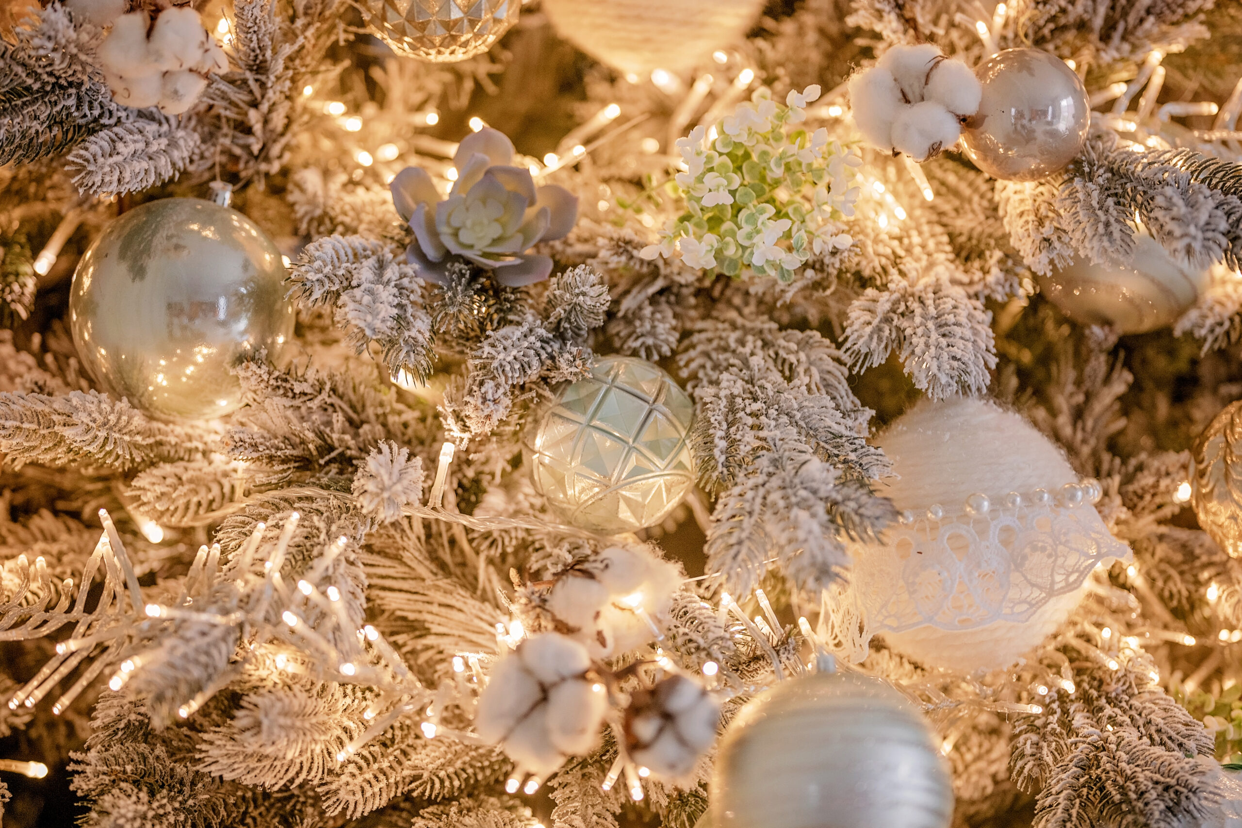 Close-up of an award-worthy Christmas tree decorated with white and silver ornaments, faux flowers, cotton, and illuminated by warm white string lights.