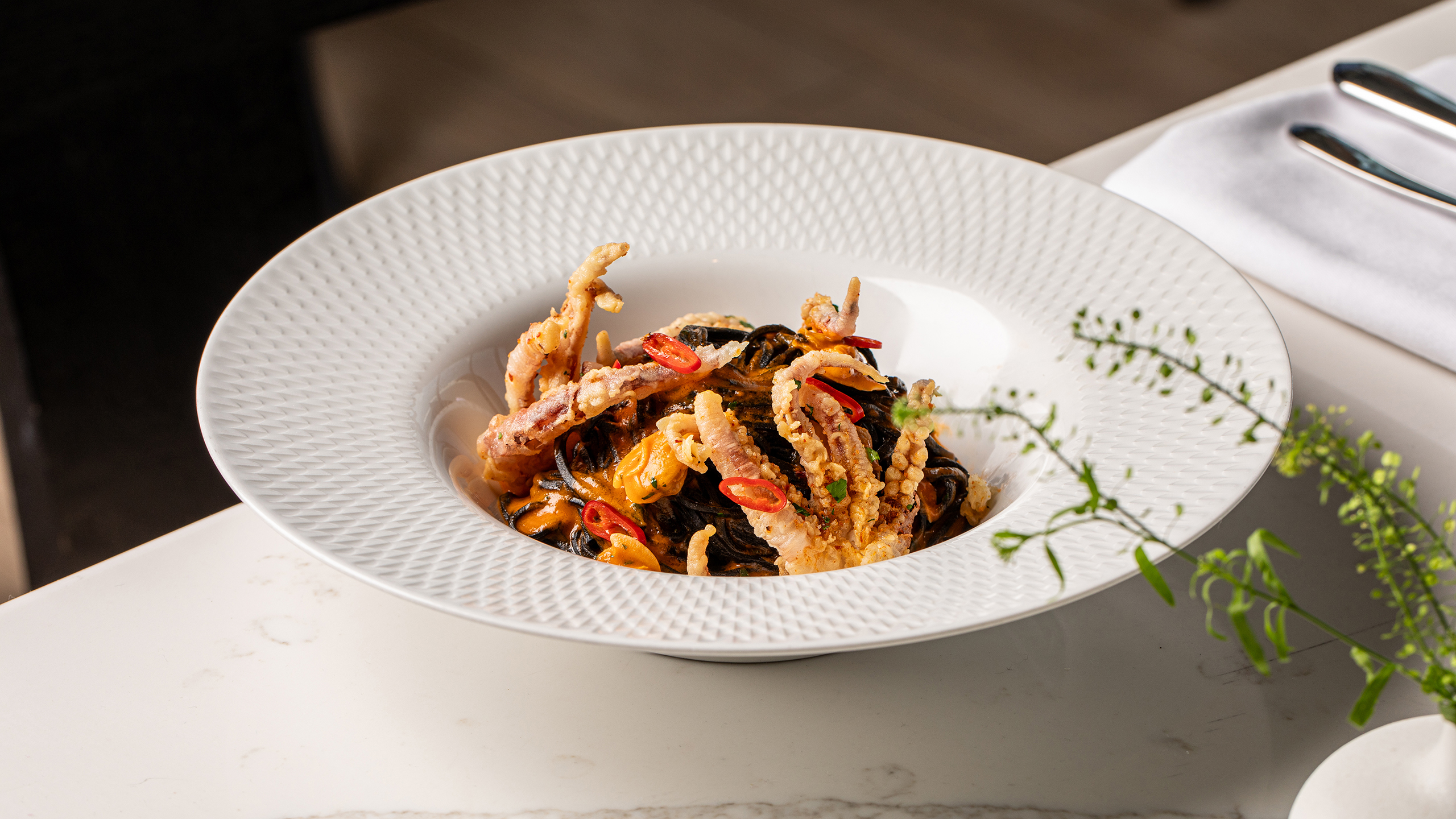 A white bowl contains pasta topped with pieces of fried soft-shell crab, sliced red chili, and green herbs, placed on a white table next to a napkin and cutlery.