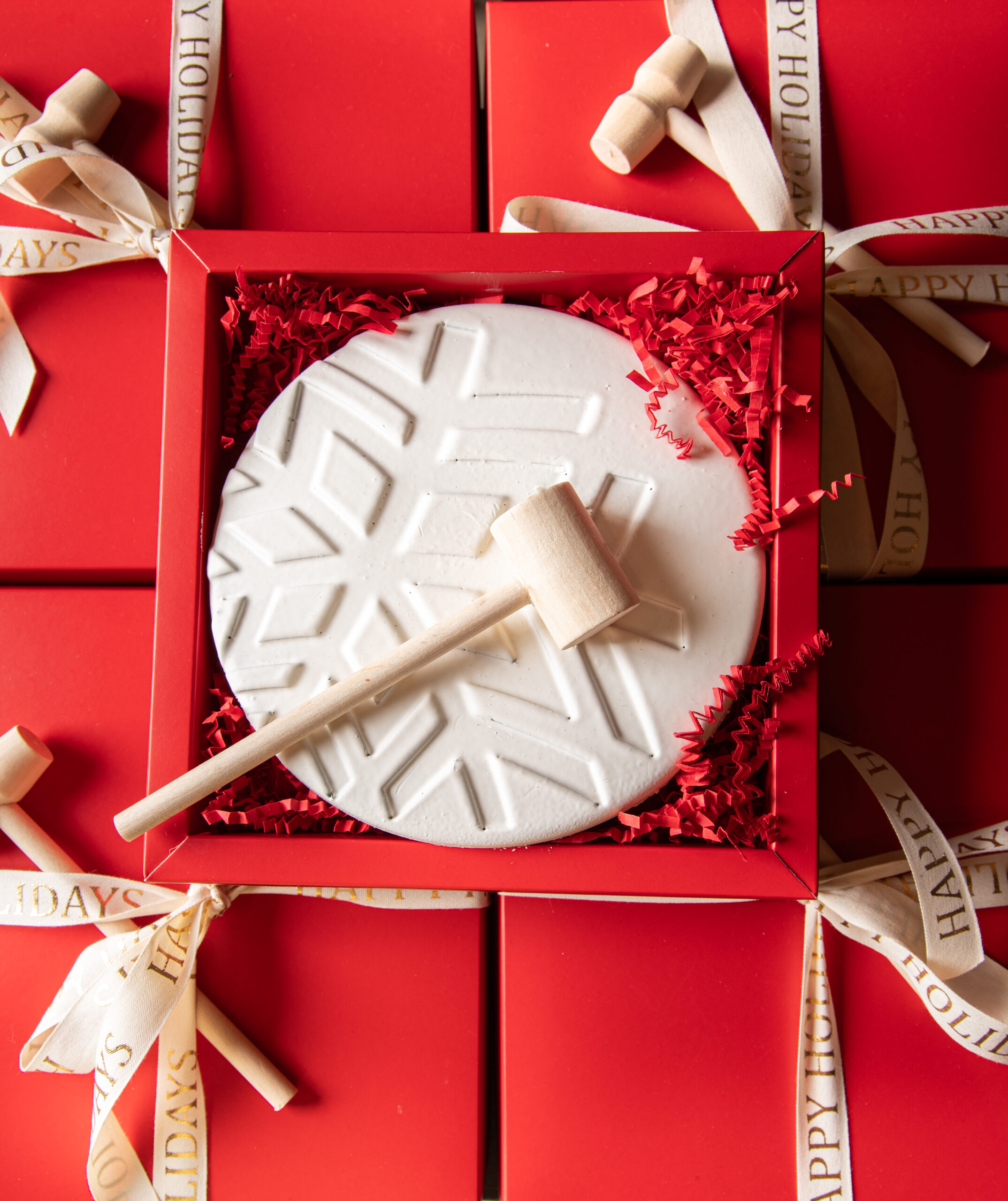 A white chocolate disk with a snowflake design and a small wooden mallet in a red gift box, surrounded by red crinkle paper and “Happy Holidays” ribbons.