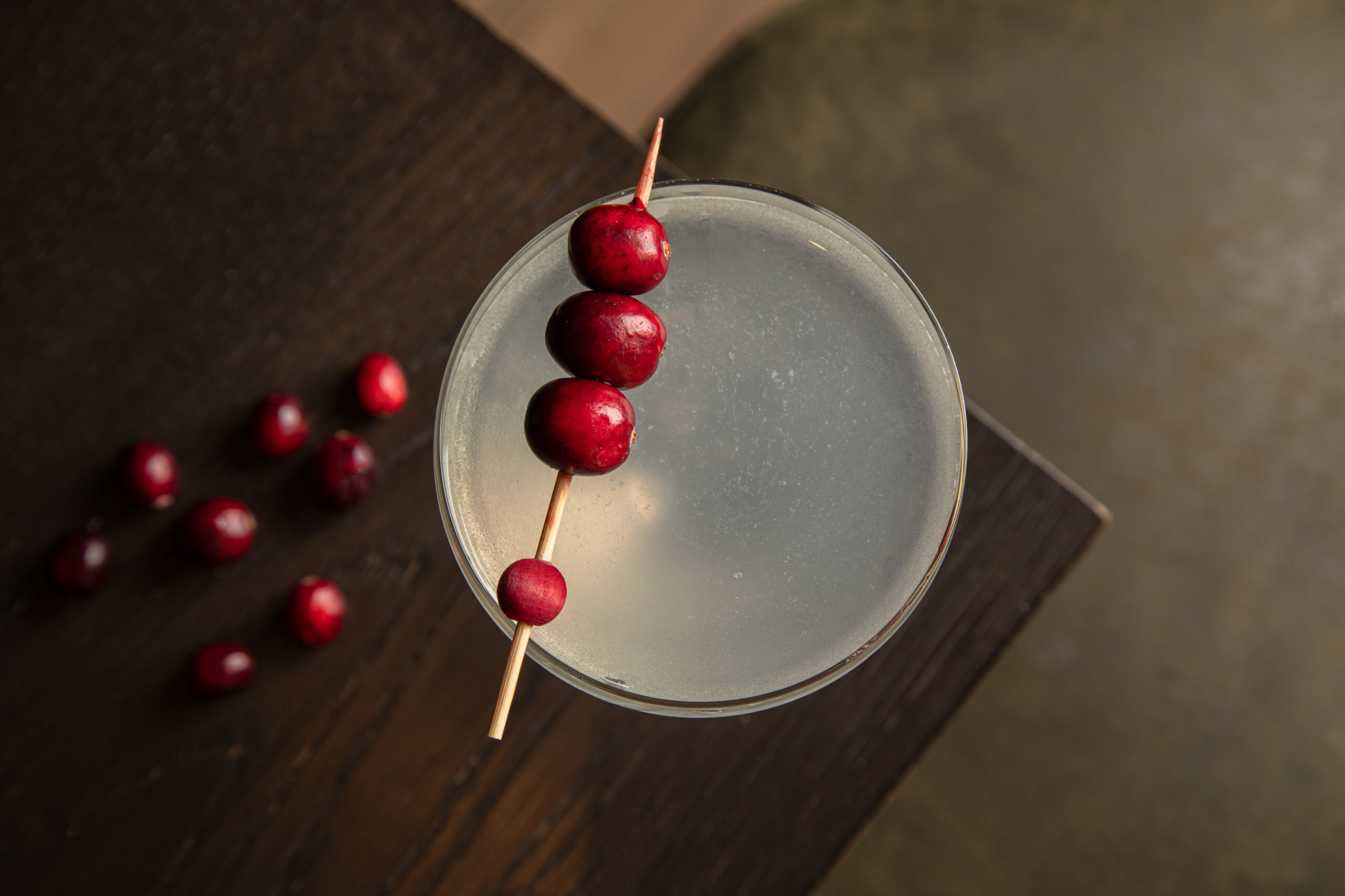 A clear cocktail in a martini glass garnished with a wooden skewer of cranberries, placed on a dark wooden surface with loose cranberries nearby.
