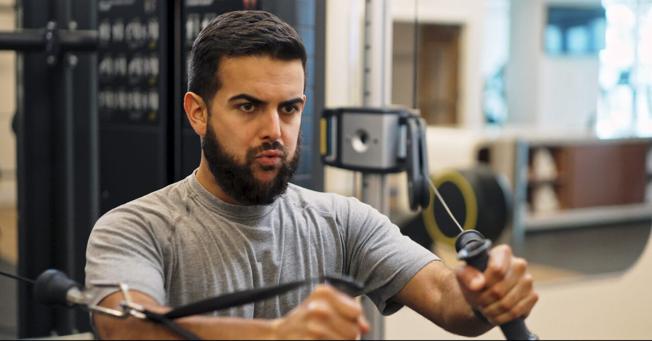A man in a gray t-shirt uses a cable machine for strength training at a gym, focusing intently during the exercise.