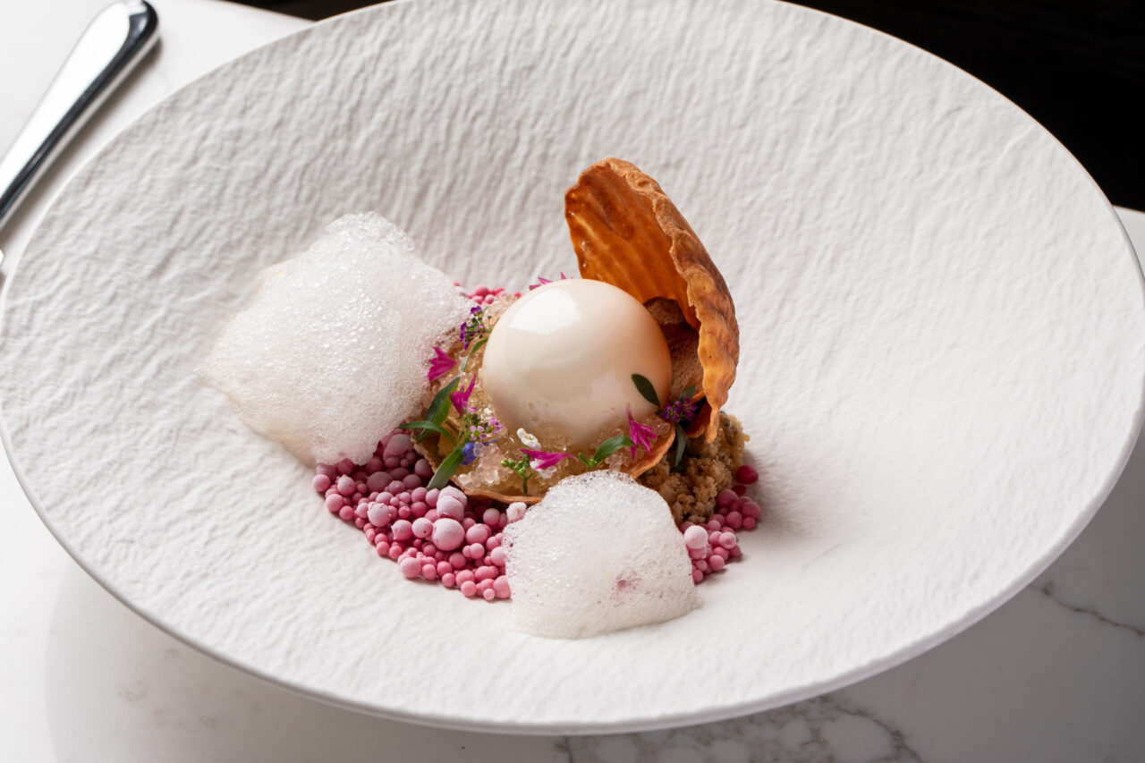 A plated dessert features a glossy white sphere, pink crunchy pearls, foam, edible flowers, a crisp wafer, and crumbly topping, all arranged on a textured white dish.