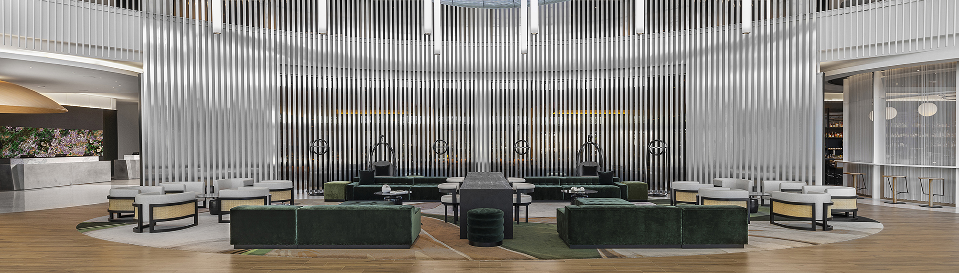 A modern hotel lobby with green velvet sofas, white armchairs, small tables, and vertical metal rods as a decorative backdrop on a circular patterned carpet.