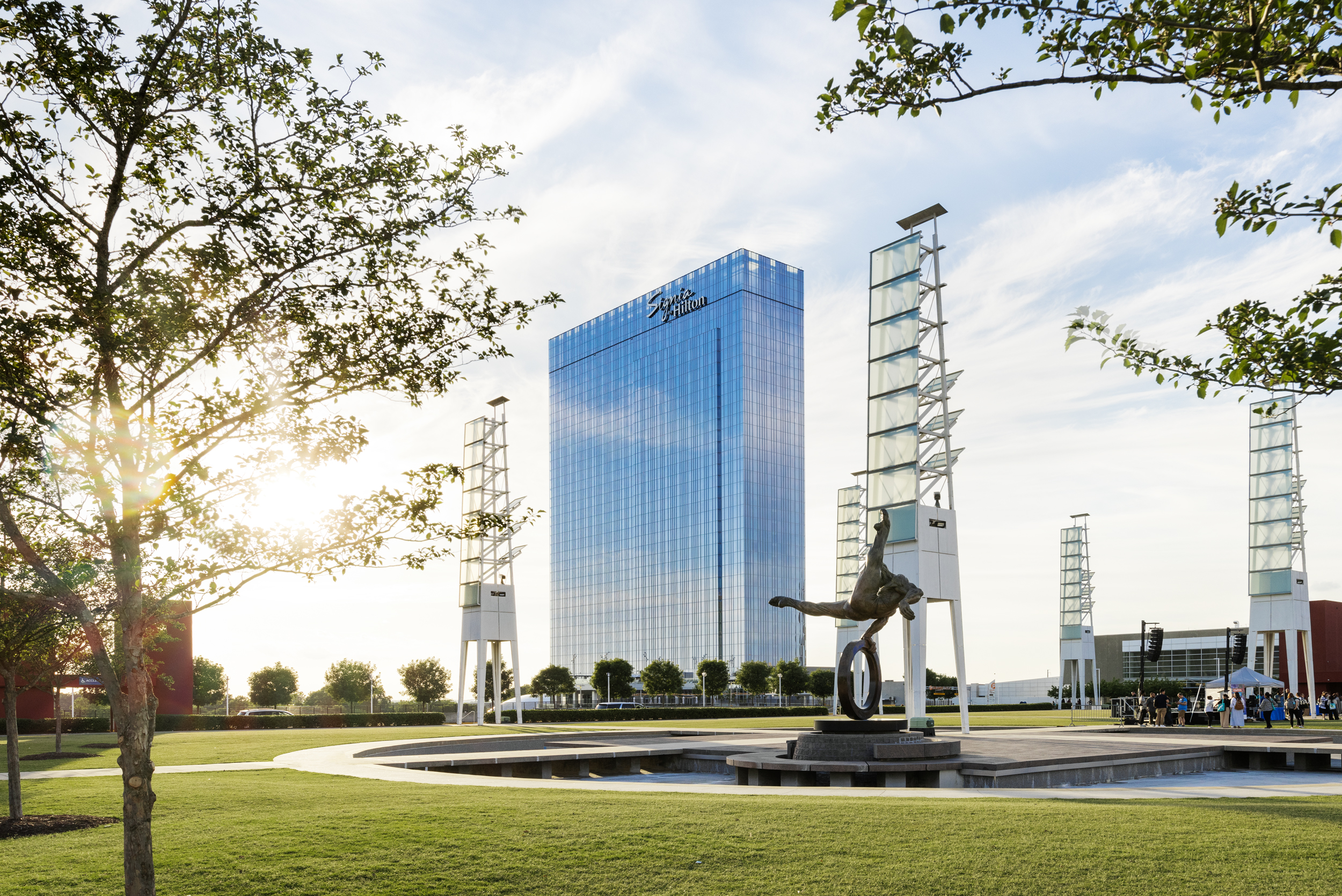 A tall glass building stands behind an award-winning sculpture of a gymnast and a circular fountain, surrounded by green grass, trees, and modern light towers.