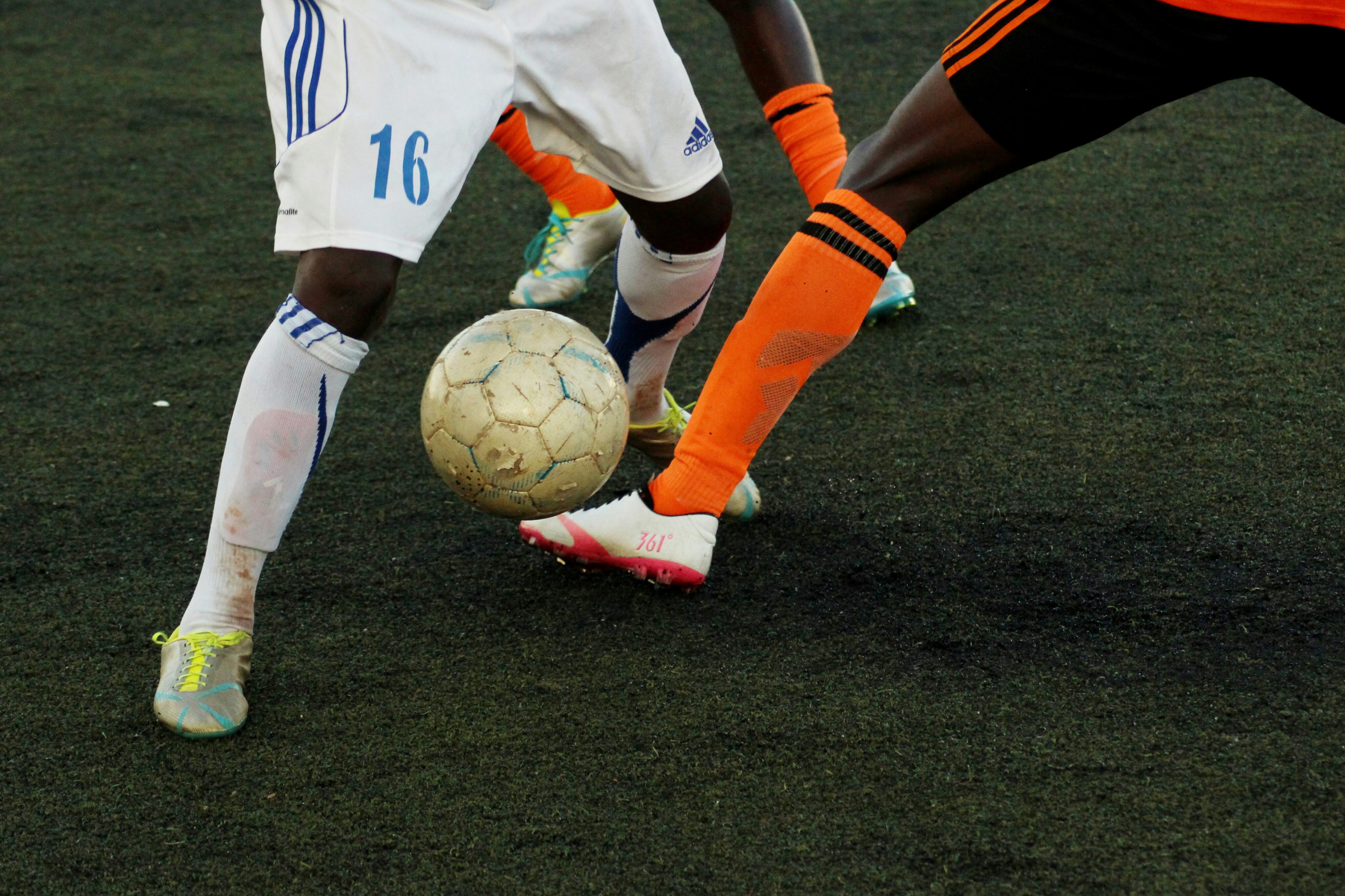 Two soccer players, one in white and one in orange, contest for the ball on a dark field. Only their legs and the ball are visible.