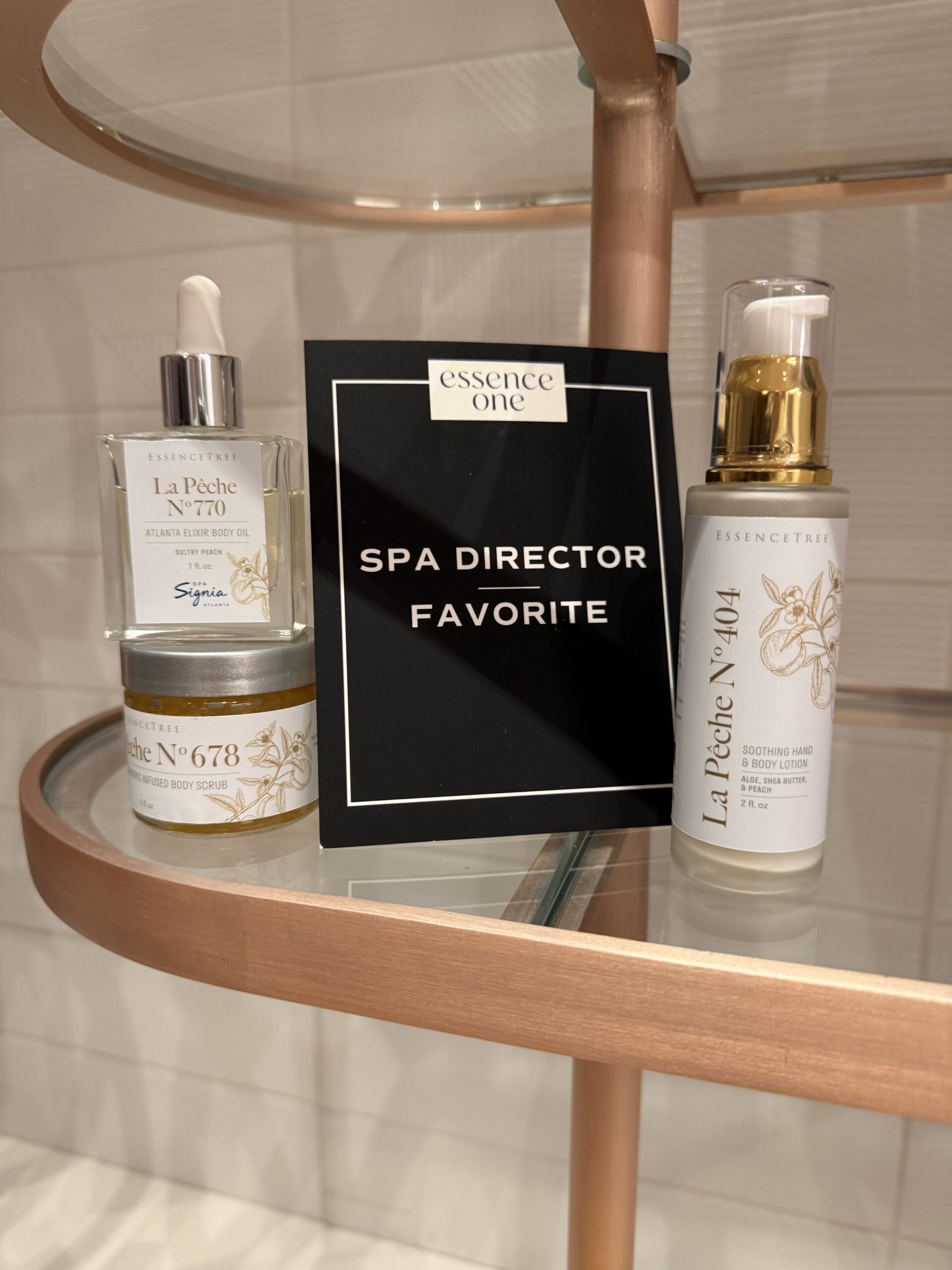 Three Essence One spa products, including a spray, a lotion, and a jar, are displayed on a glass shelf with a sign labeled “Spa Director Favorite,” highlighting each as an exceptional Spa Product.