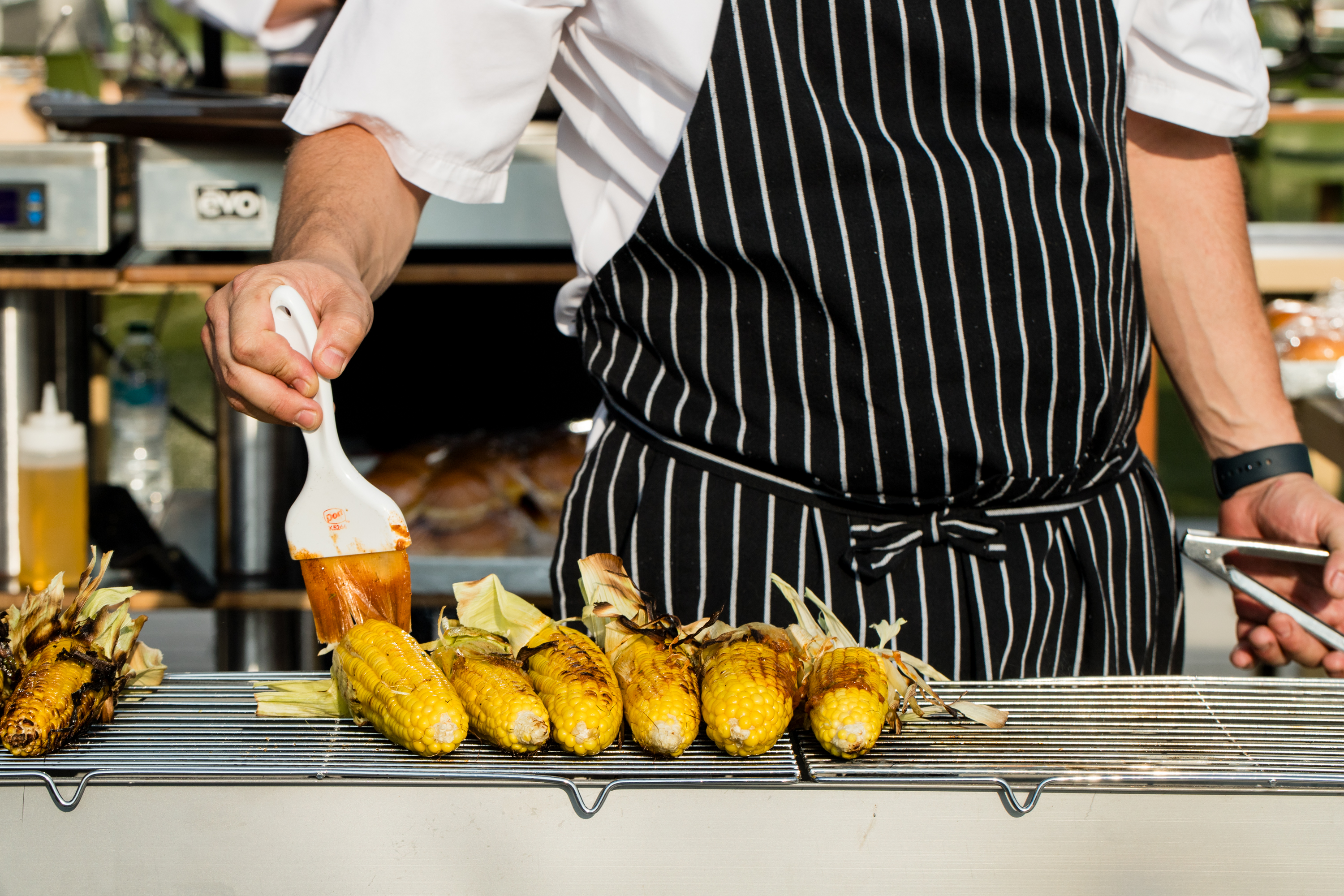 A person in a striped apron brushes sauce onto grilled corn on the cob, which is arranged on a metal rack.