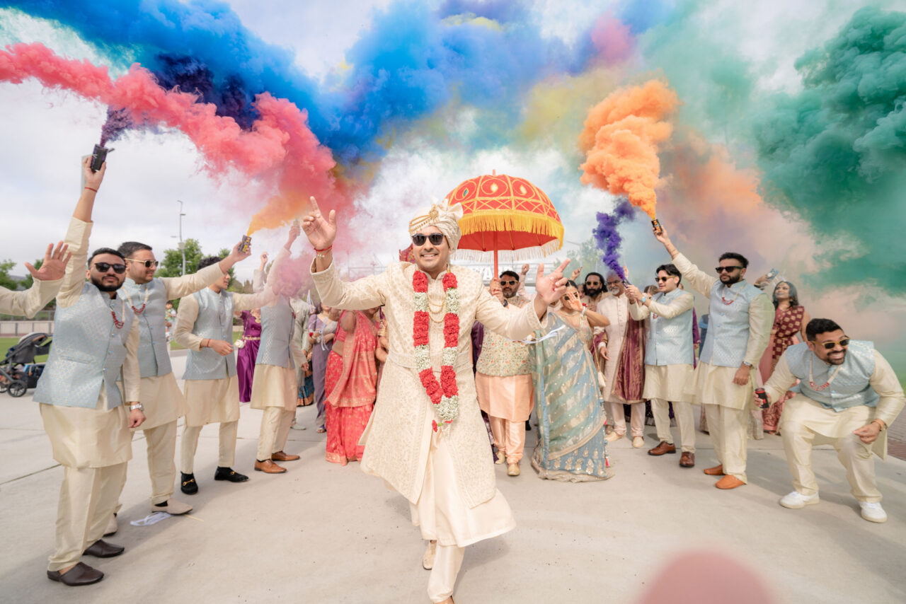 A groom in traditional attire leads a wedding procession while guests hold colorful smoke sticks, creating a vibrant and festive atmosphere.