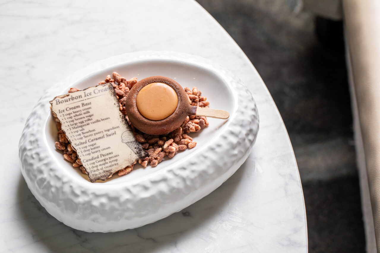 A chocolate dessert with a circular caramel center on a stick, served on puffed rice with a printed menu card, placed in a textured white dish on a marble table.